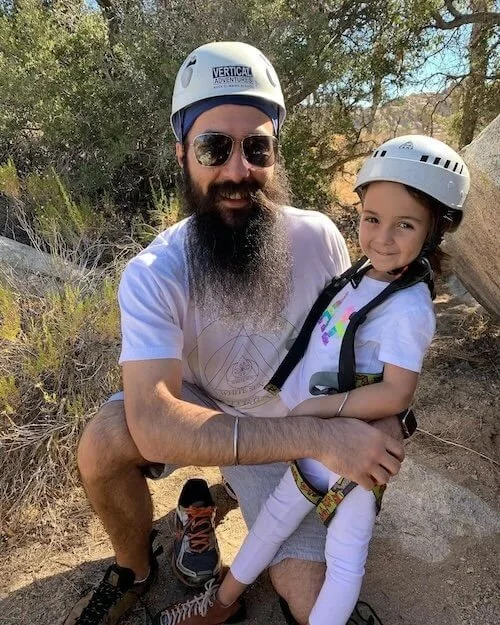 A man with a beard and sunglasses, wearing a safety helmet, crouches next to a young girl also wearing a safety helmet. They are outdoors in a natural setting with trees and rocks, suggesting they are on a hiking or adventure trip.