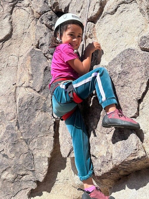 A young girl rock climbing on a rock wall, wearing a white helmet, pink shirt, and blue pants.