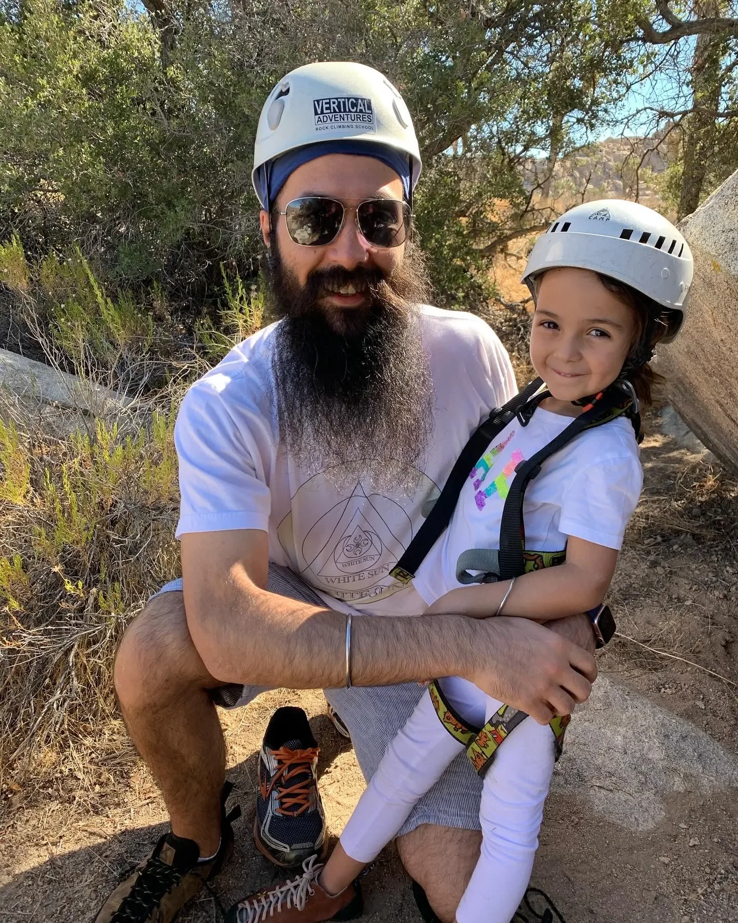 A man and a young girl wearing climbing helmets outdoors, smiling. The man has a beard and sunglasses, and the girl is holding onto the man's arm. They are on a rocky trail surrounded by trees.
