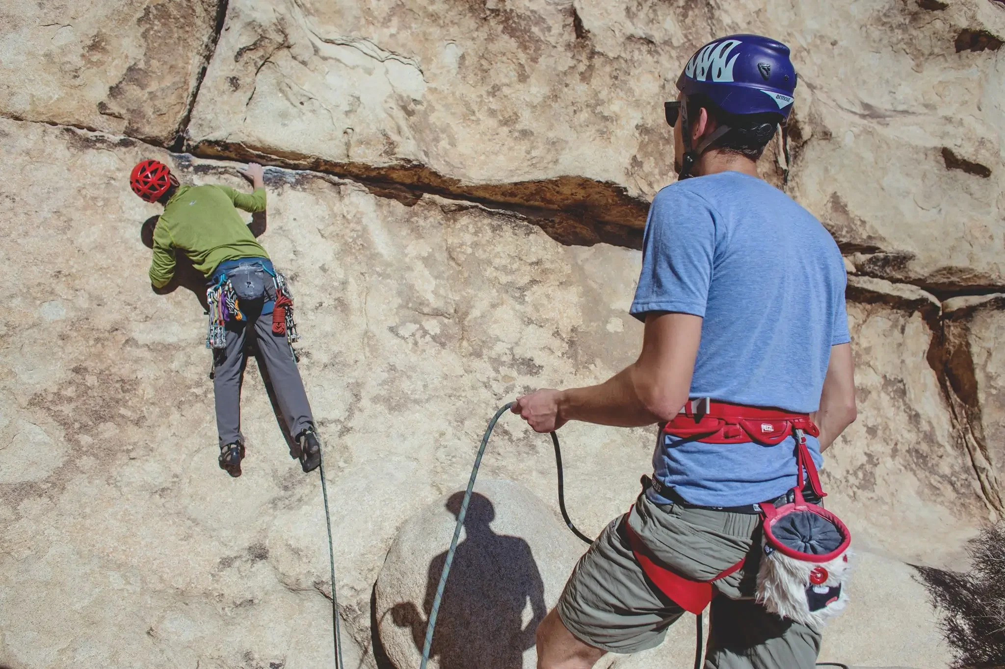 A person climbing a rock wall outdoors with a safety harness, while another person belays from below, both wearing helmets.