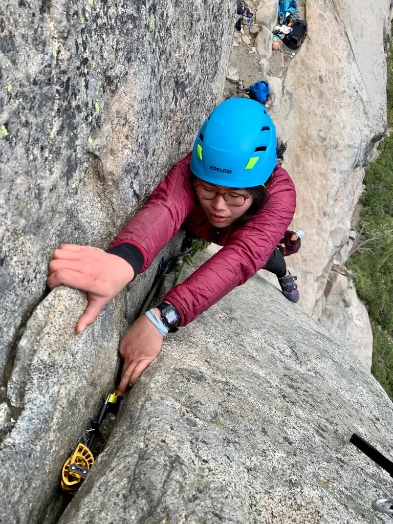 A man rock climbing on a vertical rock face, wearing a helmet, sunglasses, and climbing gear.