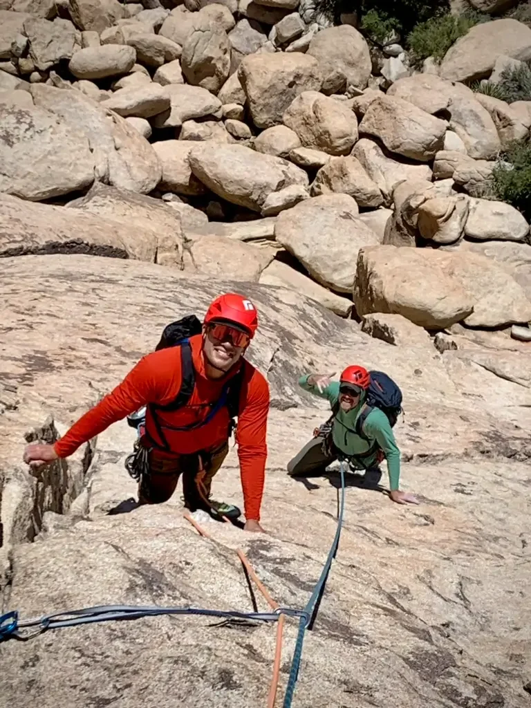 Two male rock climbers wearing red helmets and backpacks climbing on a steep rocky surface in a desert or mountainous landscape, with large boulders in the background.