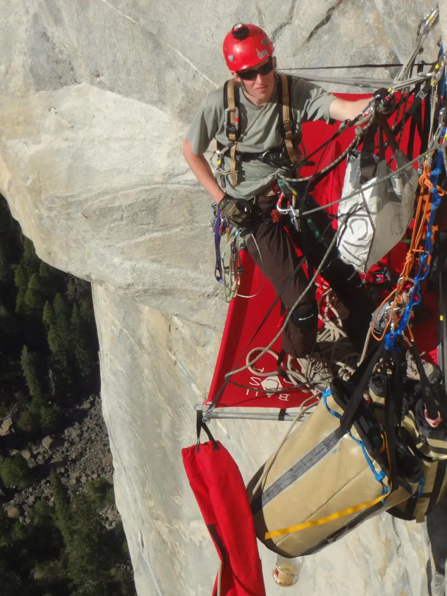 A person in climbing gear, including a red helmet and harness, sitting on a ledge of a rock face, surrounded by ropes and equipment, with a forested landscape below.