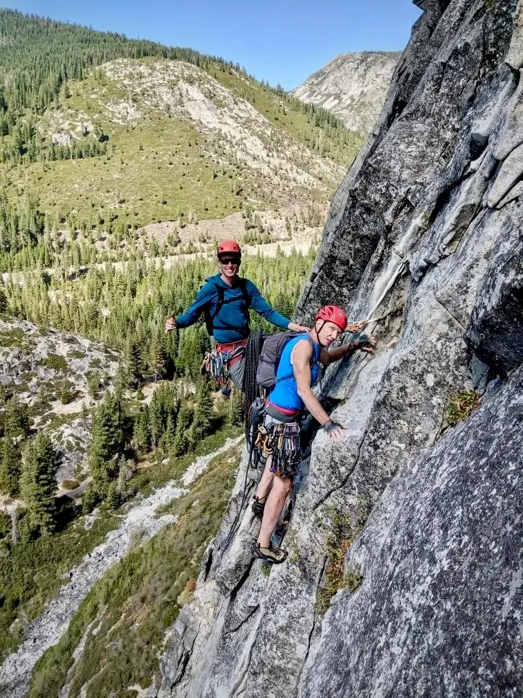 Two rock climbers, wearing helmets and harnesses, on a steep rock face outdoors with a mountain and forest in the background.