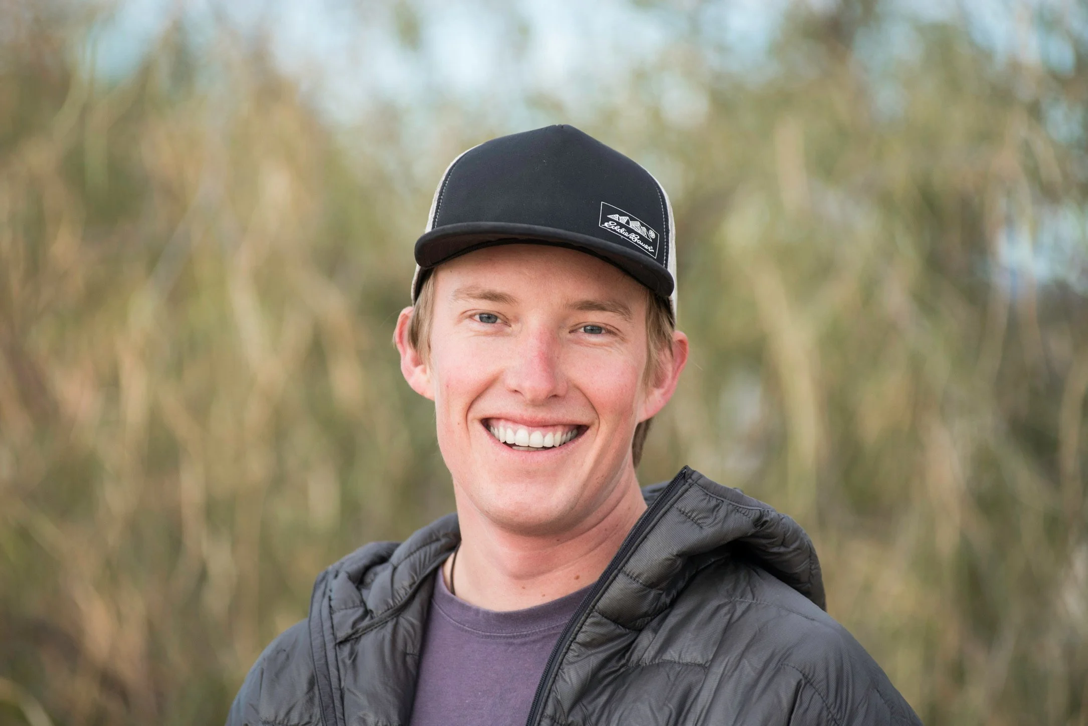 A young man smiling outdoors, wearing a black cap and a black jacket, with trees and greenery in the background.