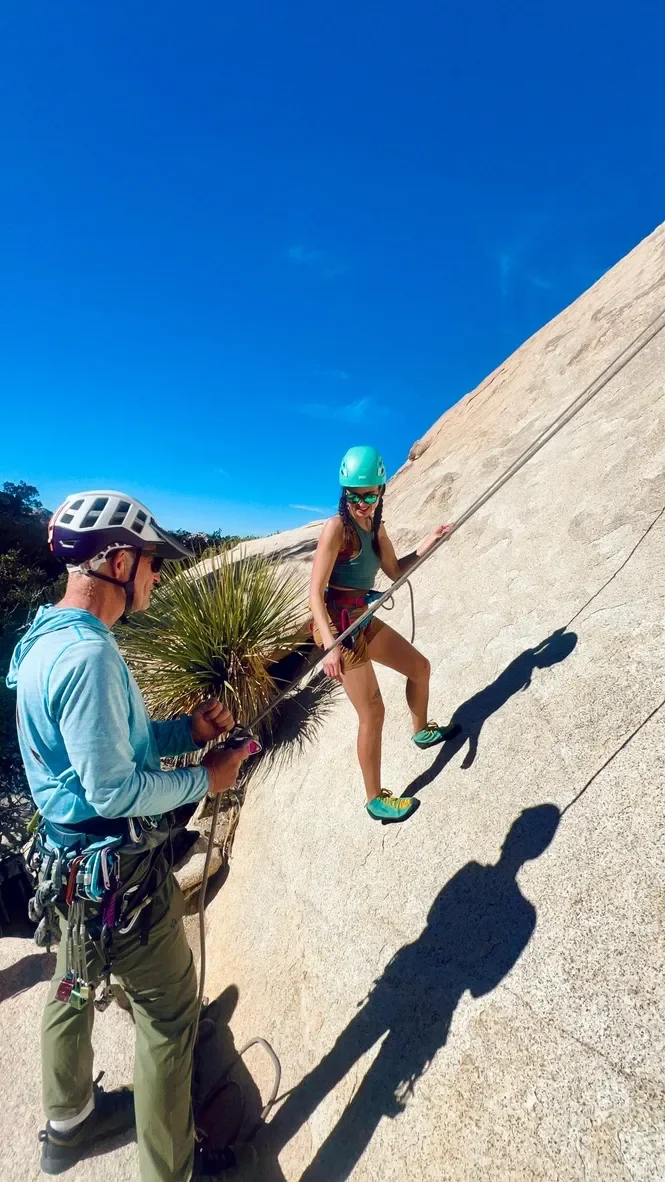 A woman rock climbing on a sloped granite surface with a man providing belay support. Both are wearing helmets and climbing gear under a clear blue sky.