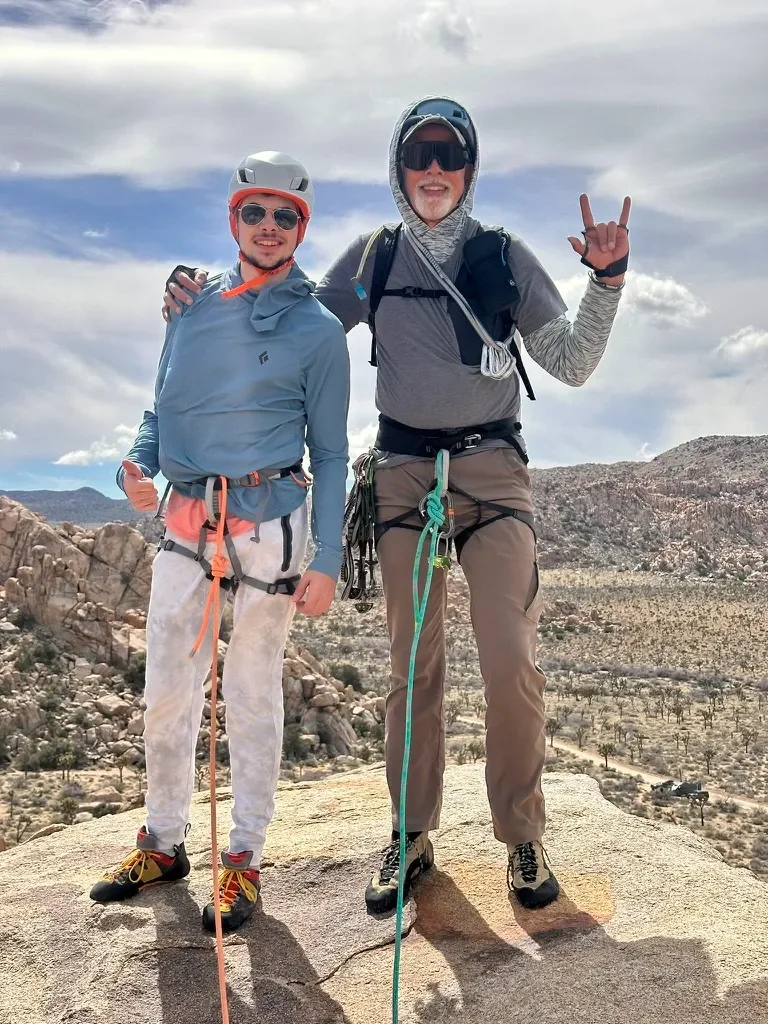 Two men dressed in climbing gear standing on a large rock formation outdoors with a mountainous desert landscape in the background. One man is making a hand gesture, and both are smiling.