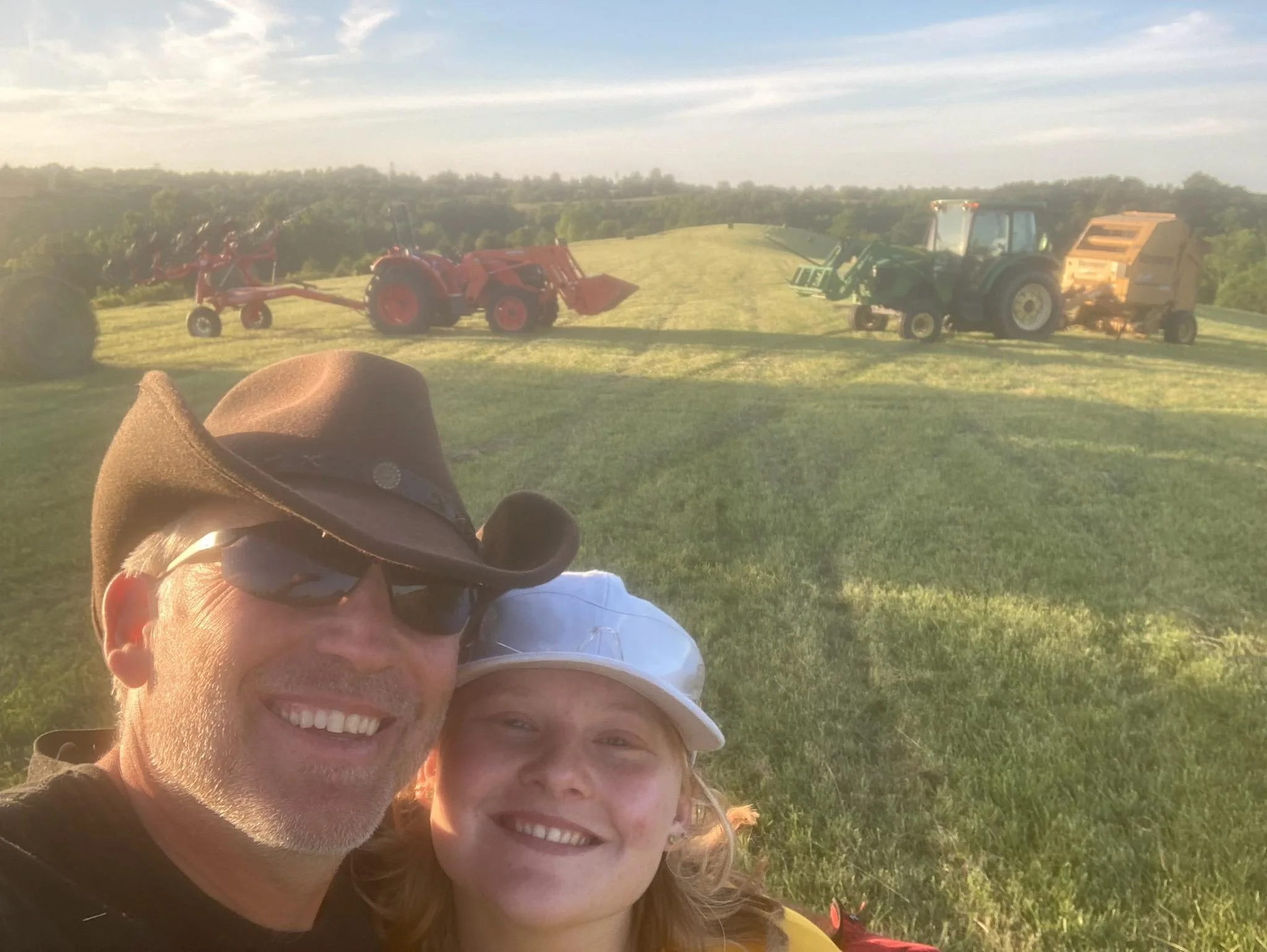 A man and a girl smiling in the foreground, with farm equipment including tractors in a field in the background, during sunset.