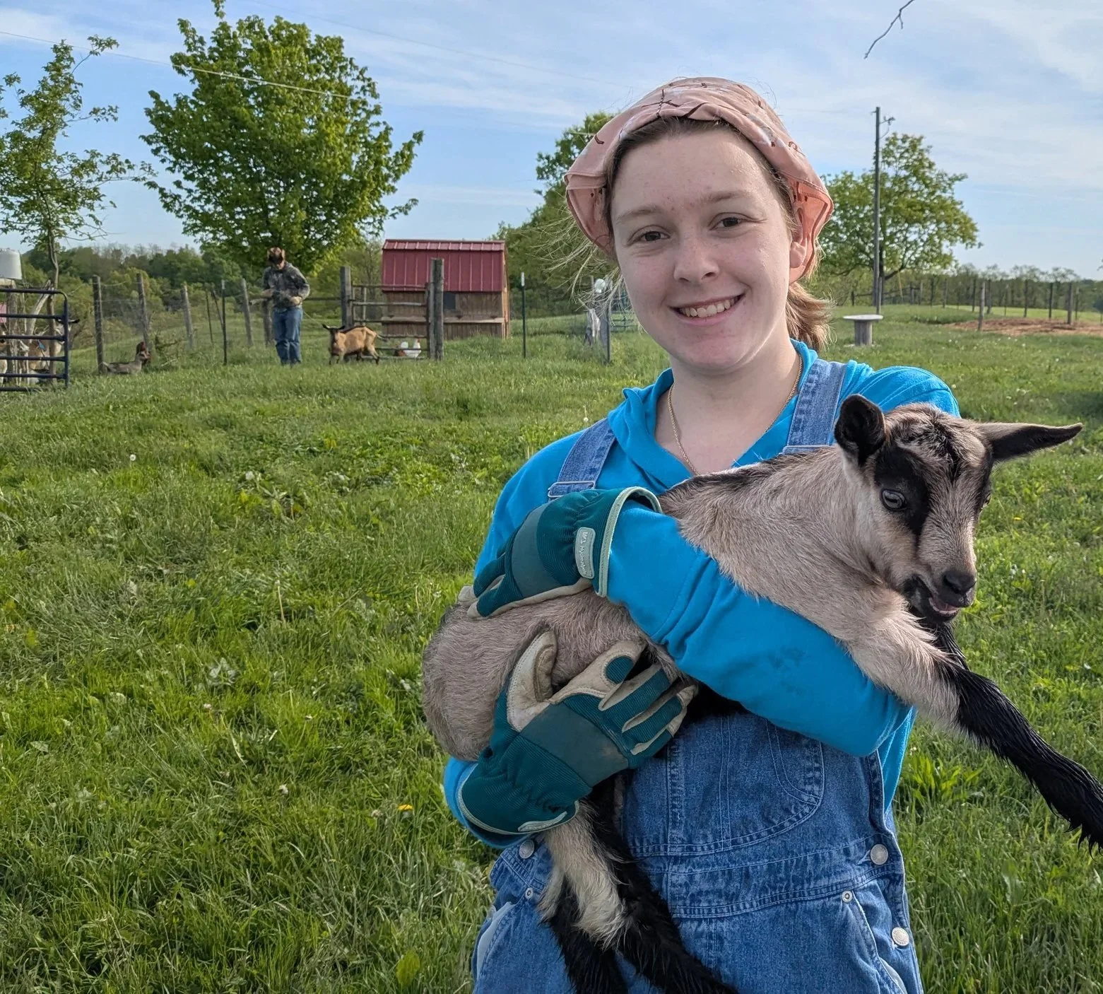 A young woman in blue overalls and gloves holding a small baby goat with black and white markings, in a grassy field with farm animals and a person in the background.
