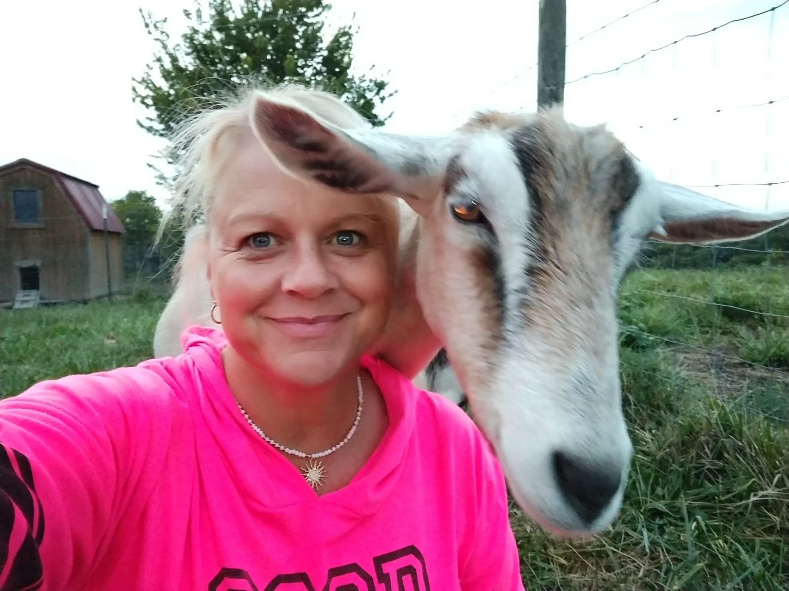 A woman wearing a pink shirt and necklace smiling with a goat beside her in a grassy outdoor area with a barn and trees in the background.