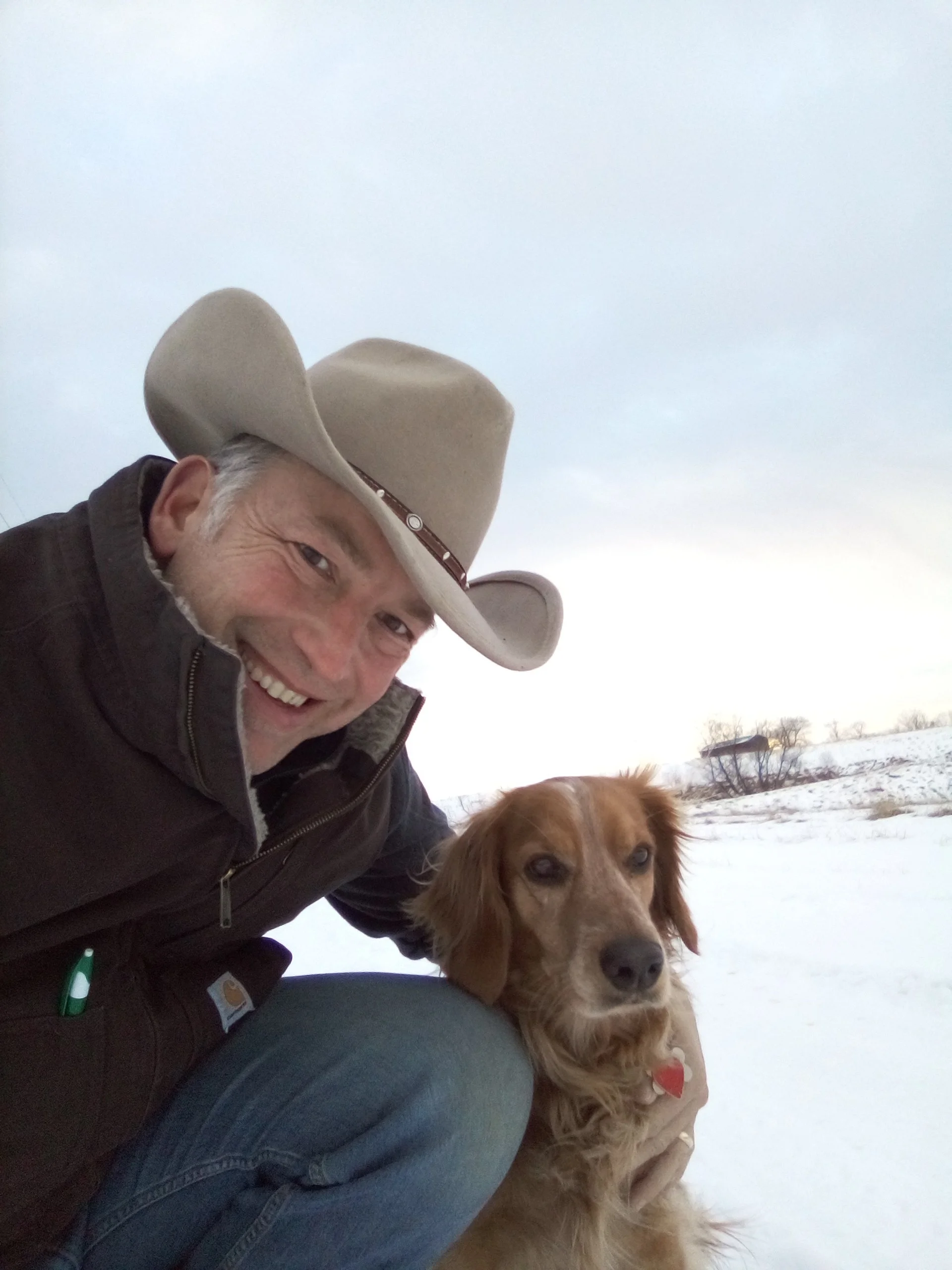 A man smiling with a golden retriever in a snowy outdoor landscape, both wearing winter clothing and the man wearing a beige cowboy hat.
