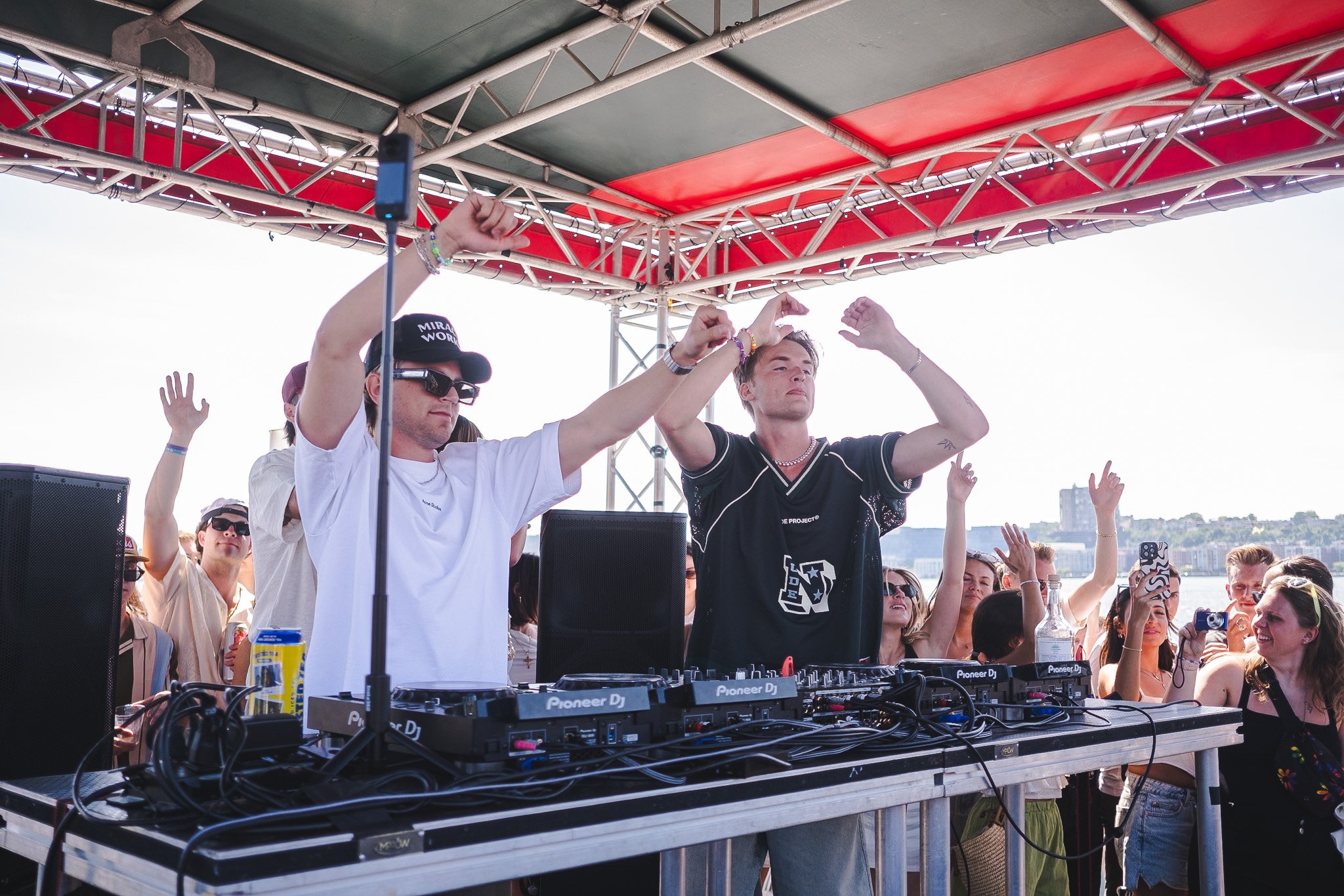 Two male DJs perform on an outdoor stage with a crowd dancing in front of them. The stage has a red canopy supported by metal scaffolding and the sky is clear.