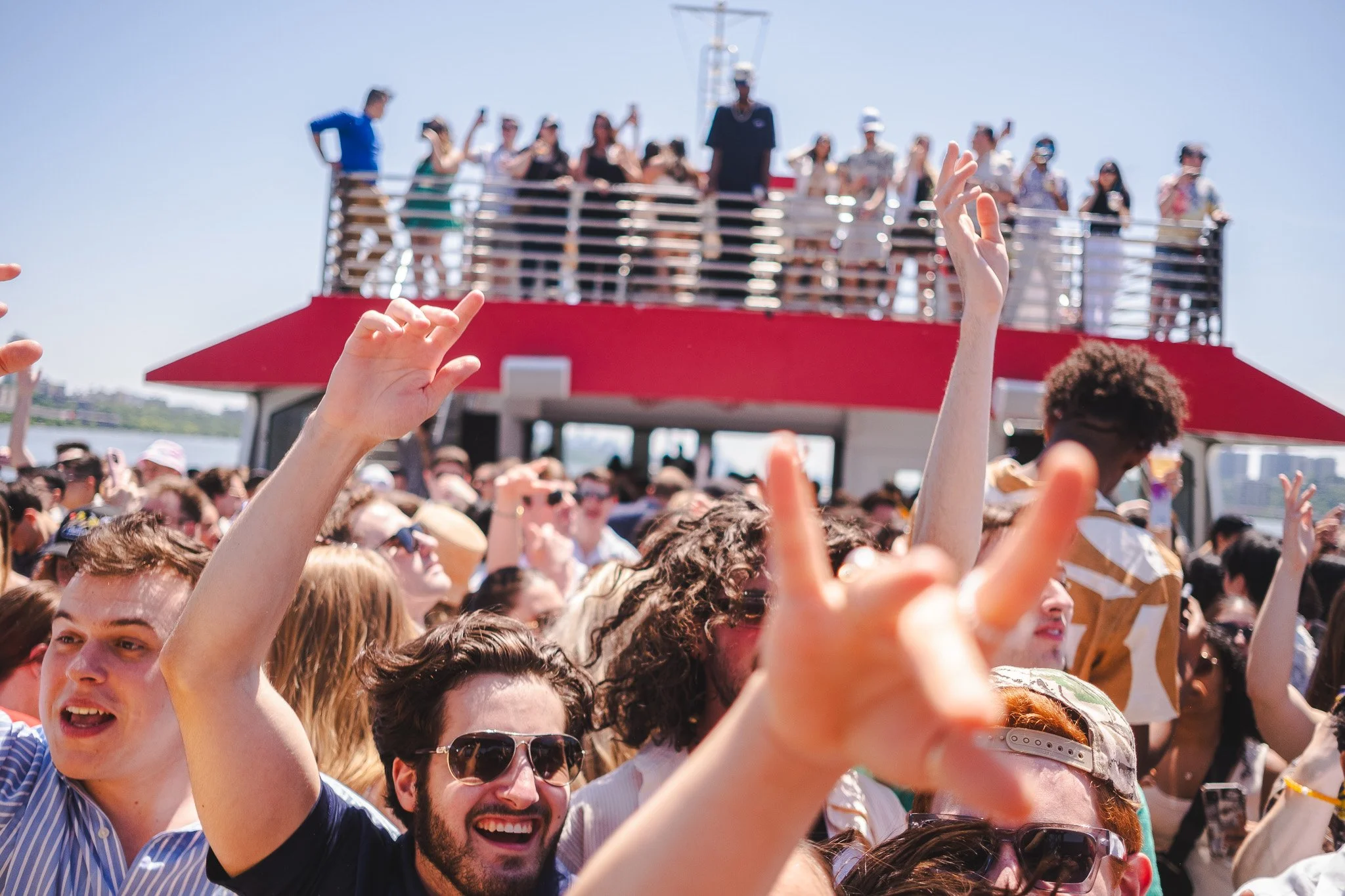 Crowd of people at an outdoor event with hands raised, and a boat or ship with people on the deck in the background under a clear blue sky.