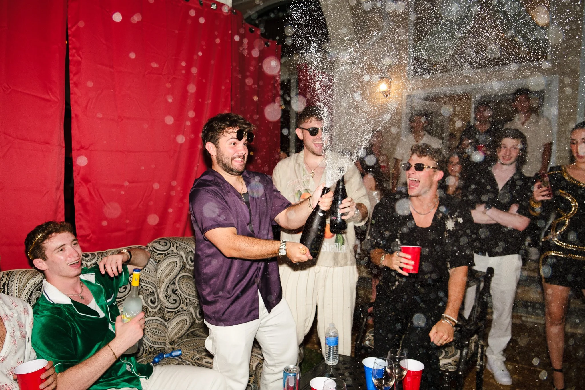 Group of young adults celebrating with champagne, some wearing sunglasses, at a party with red curtain backdrop and people holding drinks, with confetti or foam in the air.