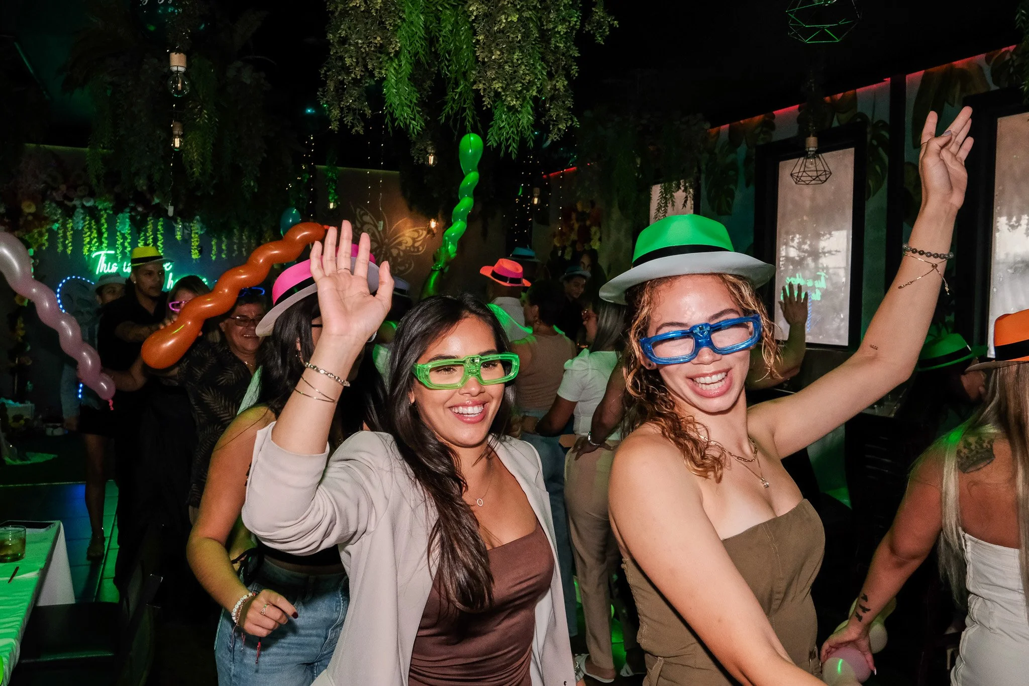 People dancing and celebrating at a party with festive hats and fun glasses, surrounded by colorful balloons and green lighting.