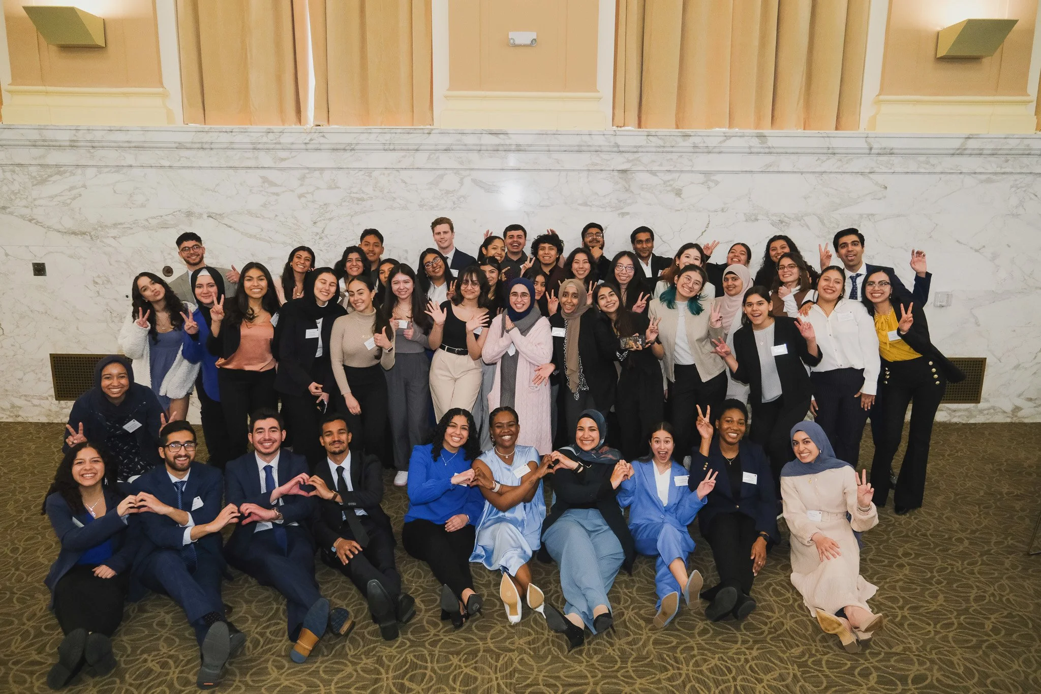 Group of diverse young adults posing together indoors, many making peace signs or heart shapes with their hands, smiling for the camera.