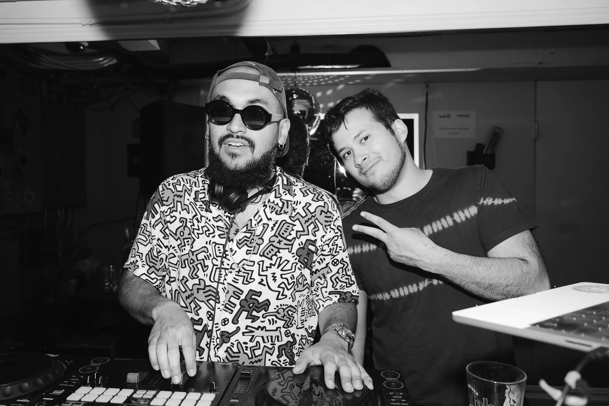 Two men, one wearing sunglasses and headphones around his neck, mixing music at a DJ turntable in a dimly lit room.