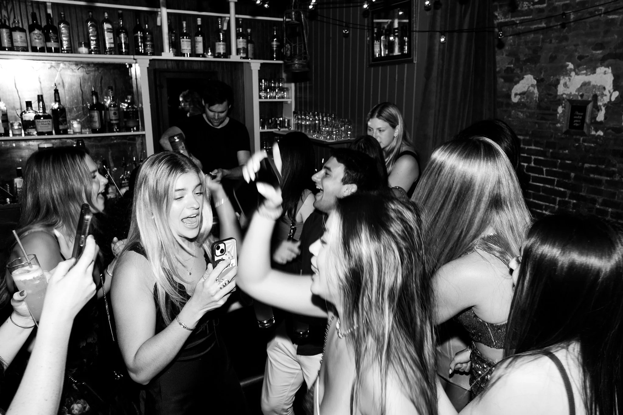 A black-and-white photo of a lively social gathering in a bar or nightclub with several women near the camera holding drinks and taking photos, and a bartender pouring drinks in the background.