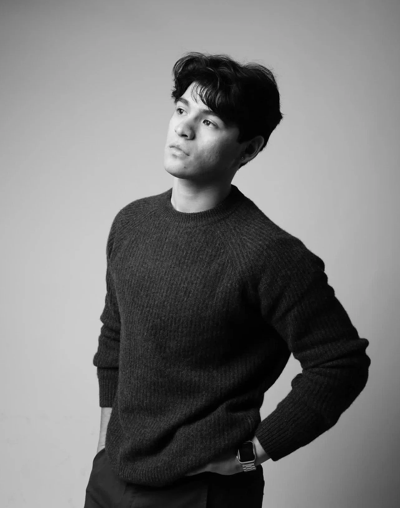 Black and white portrait of a young man with dark hair, wearing a dark sweater, looking thoughtfully to the side, with his hand on his hip, against a neutral background.