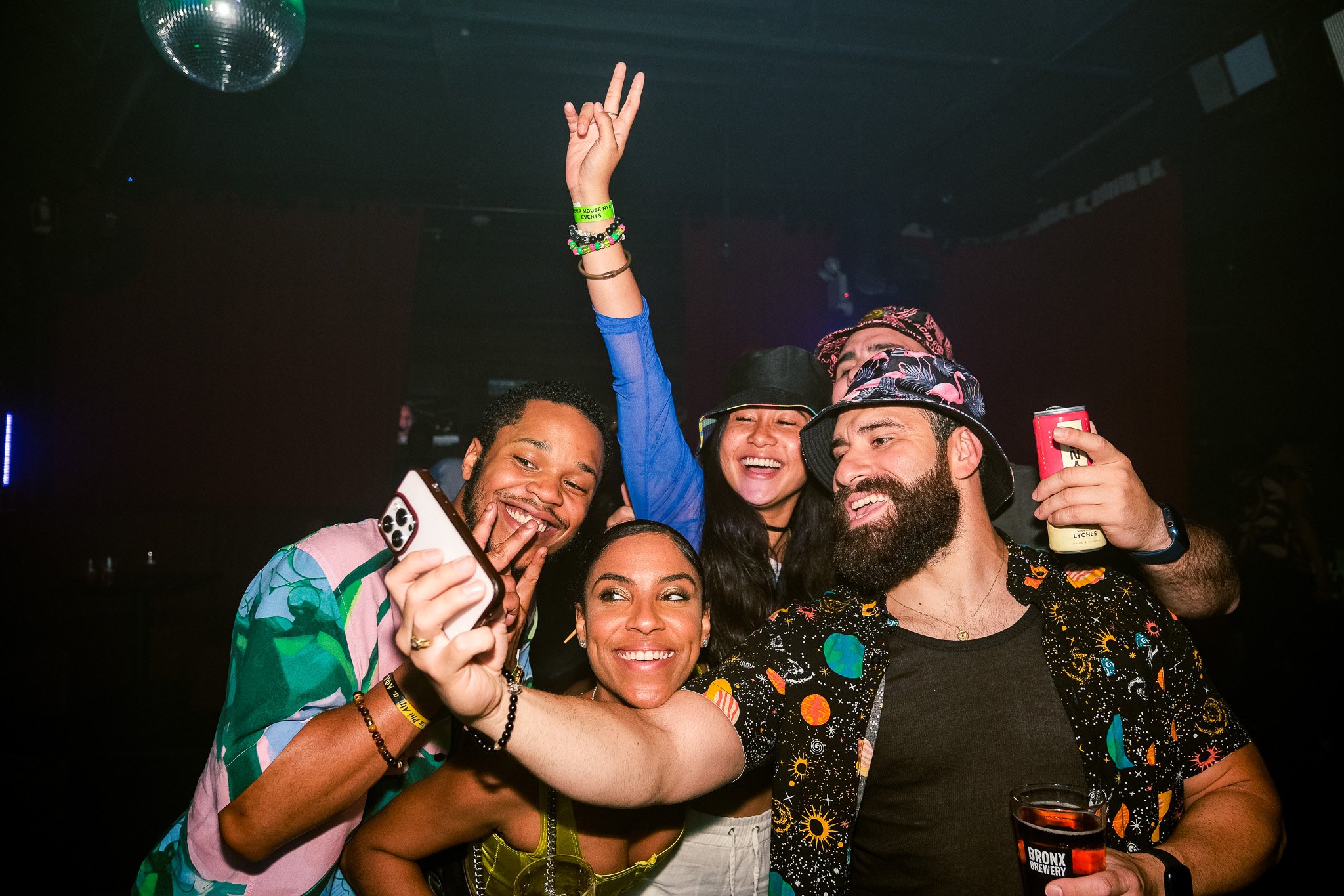 A group of six friends happily taking a selfie at a party with a dark background and a disco ball overhead. They are smiling and wearing colorful, festive clothing and accessories.