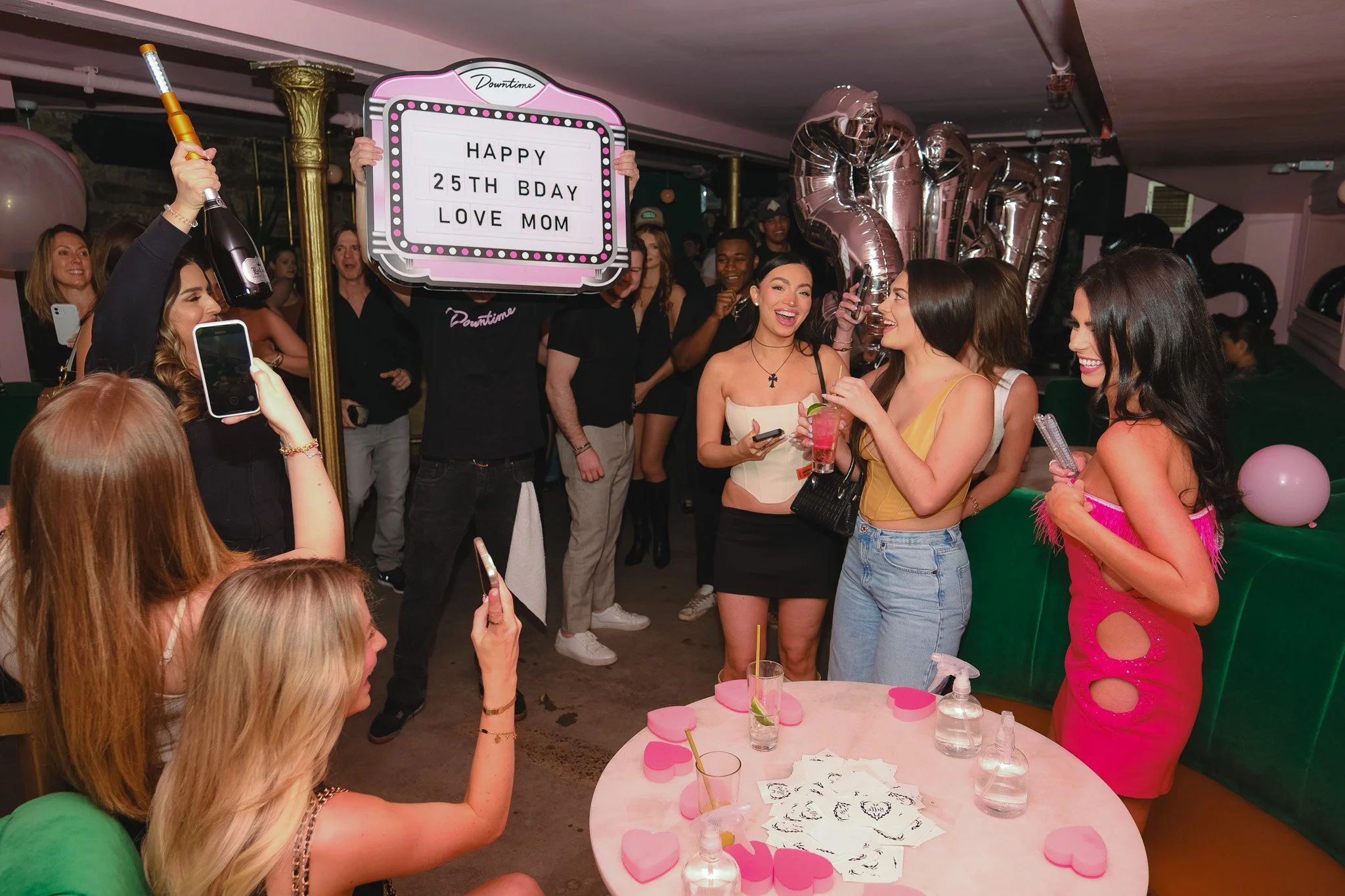 A group of women celebrating a birthday at a party, with decorations, balloons, and drinks, as someone holds a sign that reads 'HAPPY 25th BDAY LOVE MOM'.