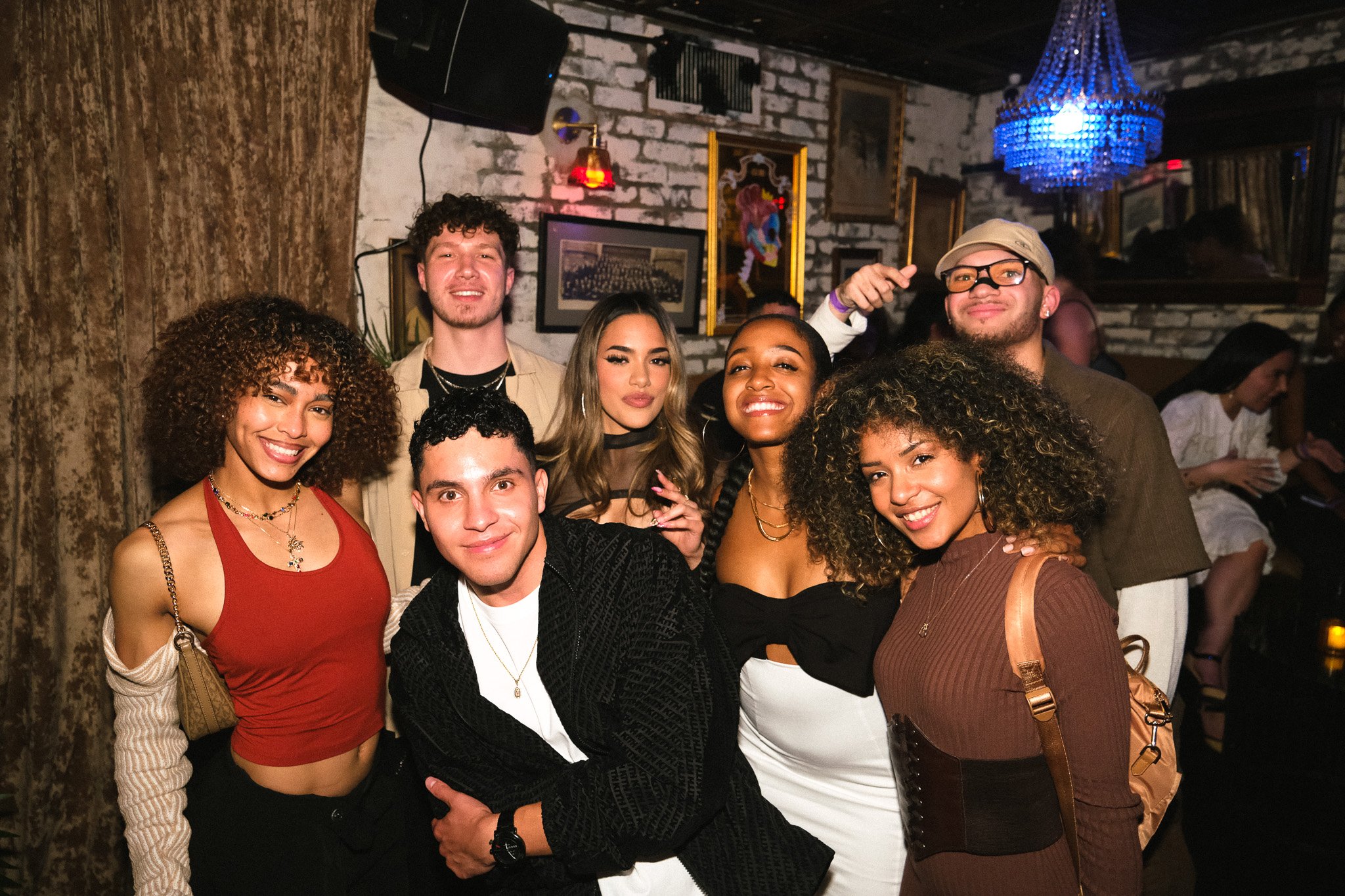 Group of diverse friends smiling and posing together at a social gathering or party in a dimly lit bar or club, with brick walls and framed artwork in the background.