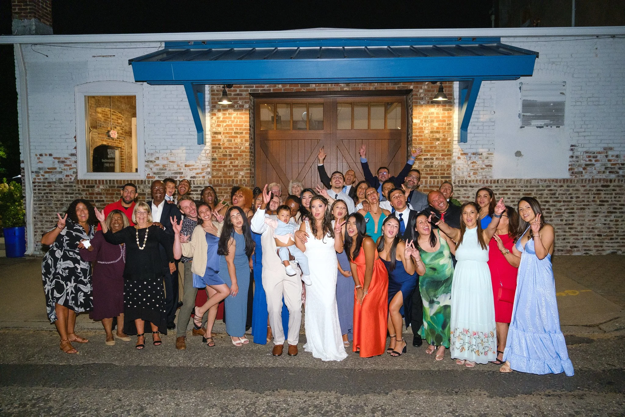 A large group of people celebrating in front of a brick building at night. They are smiling, making peace signs, and raising their hands in the air. Many are dressed in colorful, festive clothing, suggesting a party or special event.