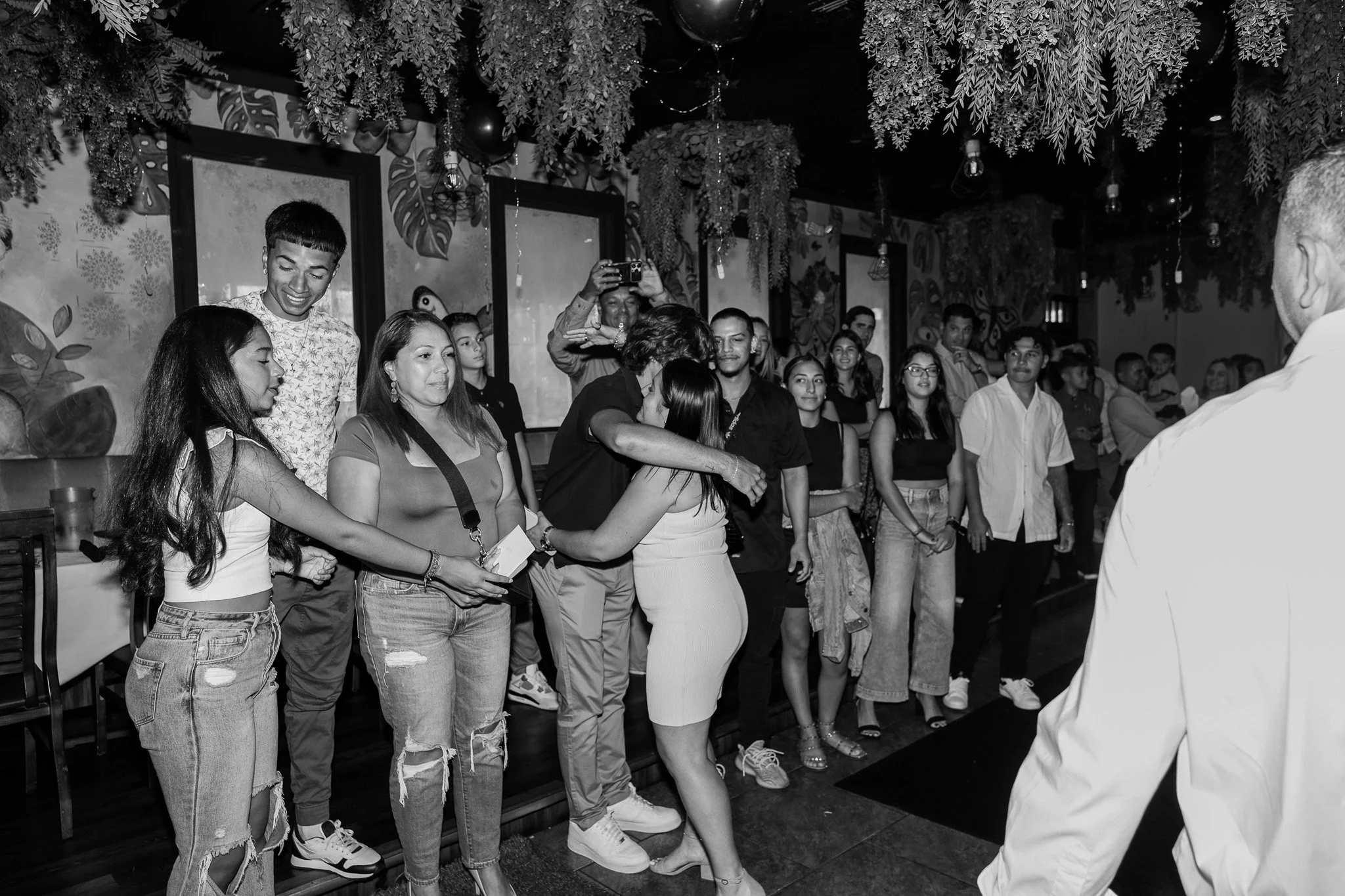 A group of people gathered indoors, some are embracing or chatting, with a mist of hanging greenery and decorative elements above them.