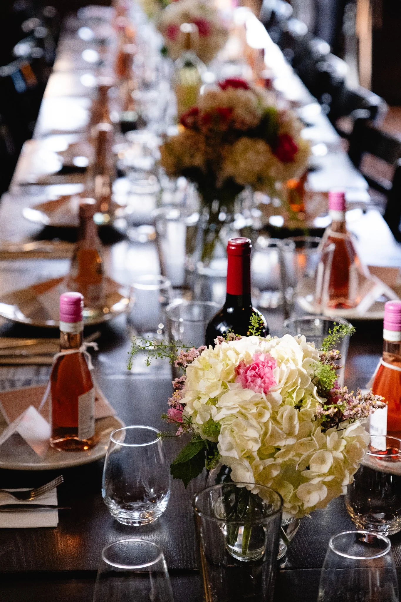 A floral centerpiece with white and pink flowers, surrounded by wine bottles, glasses, and place settings on a long dining table.