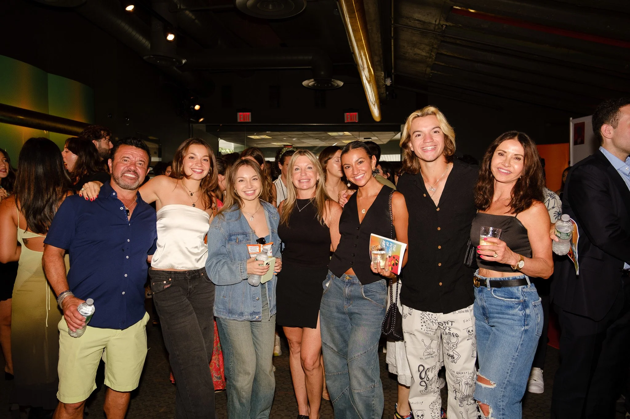 Group of diverse people posing happily for a photo at an indoor event or gathering. They are smiling and standing close together, some holding drinks or bottles of water.