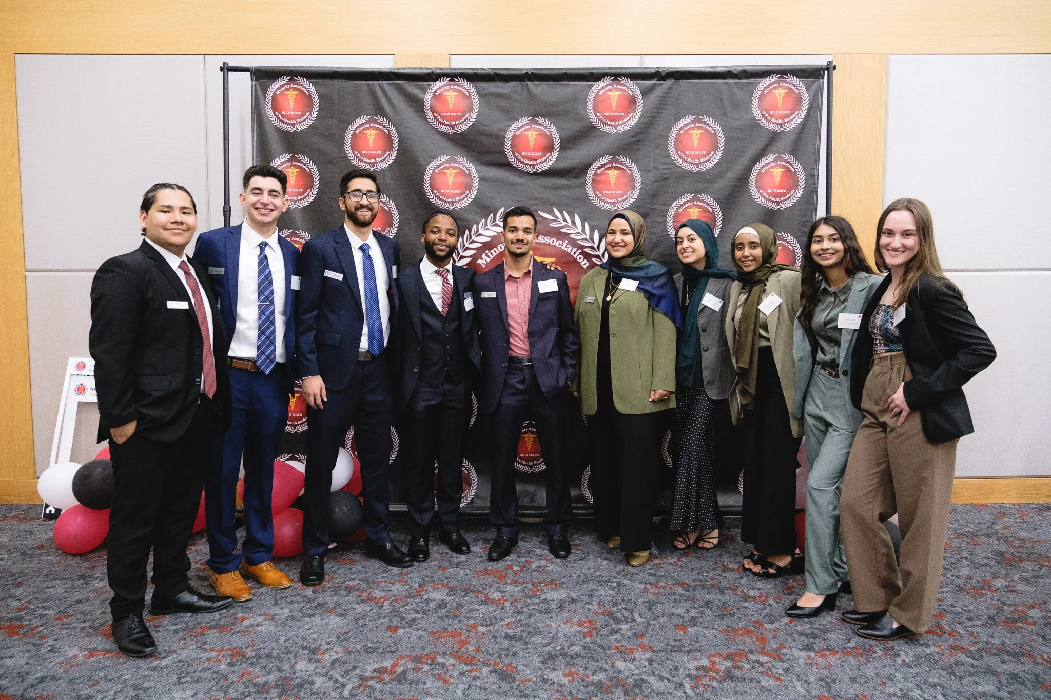 A group of nine diverse young adults standing together at an indoor event, smiling in front of a black backdrop with logos and text, and balloons on the floor.