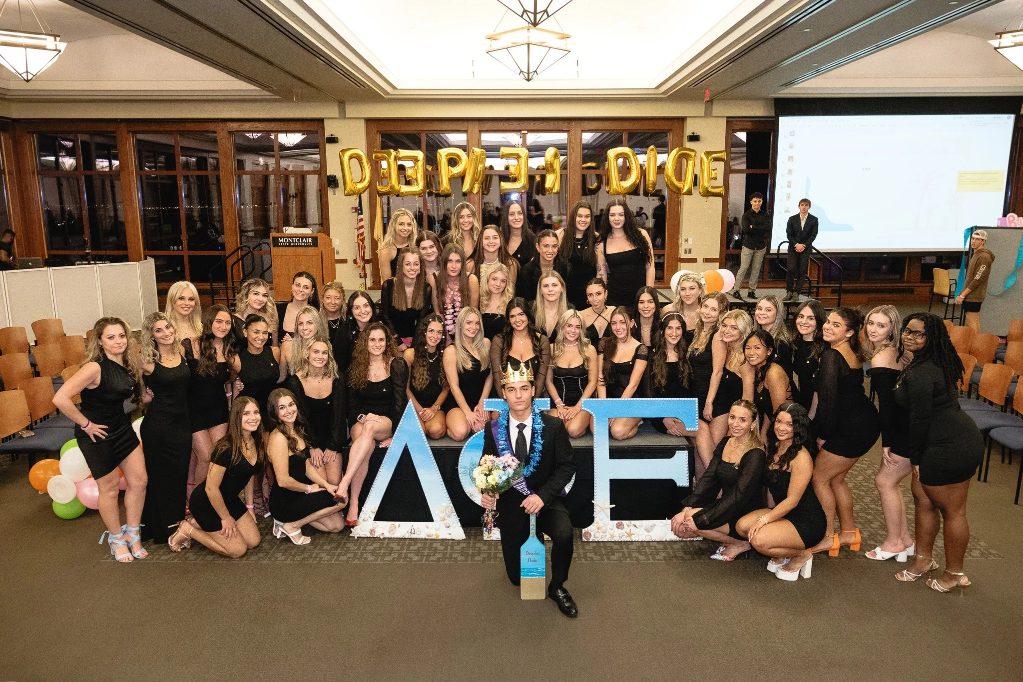 Group of young women dressed in black, gathered around a front row, during a celebration or graduation event. Decor with balloons, a large sign spelling 'DIEP' and Greek letters indicating an sorority or fraternity event. There are people in the back
