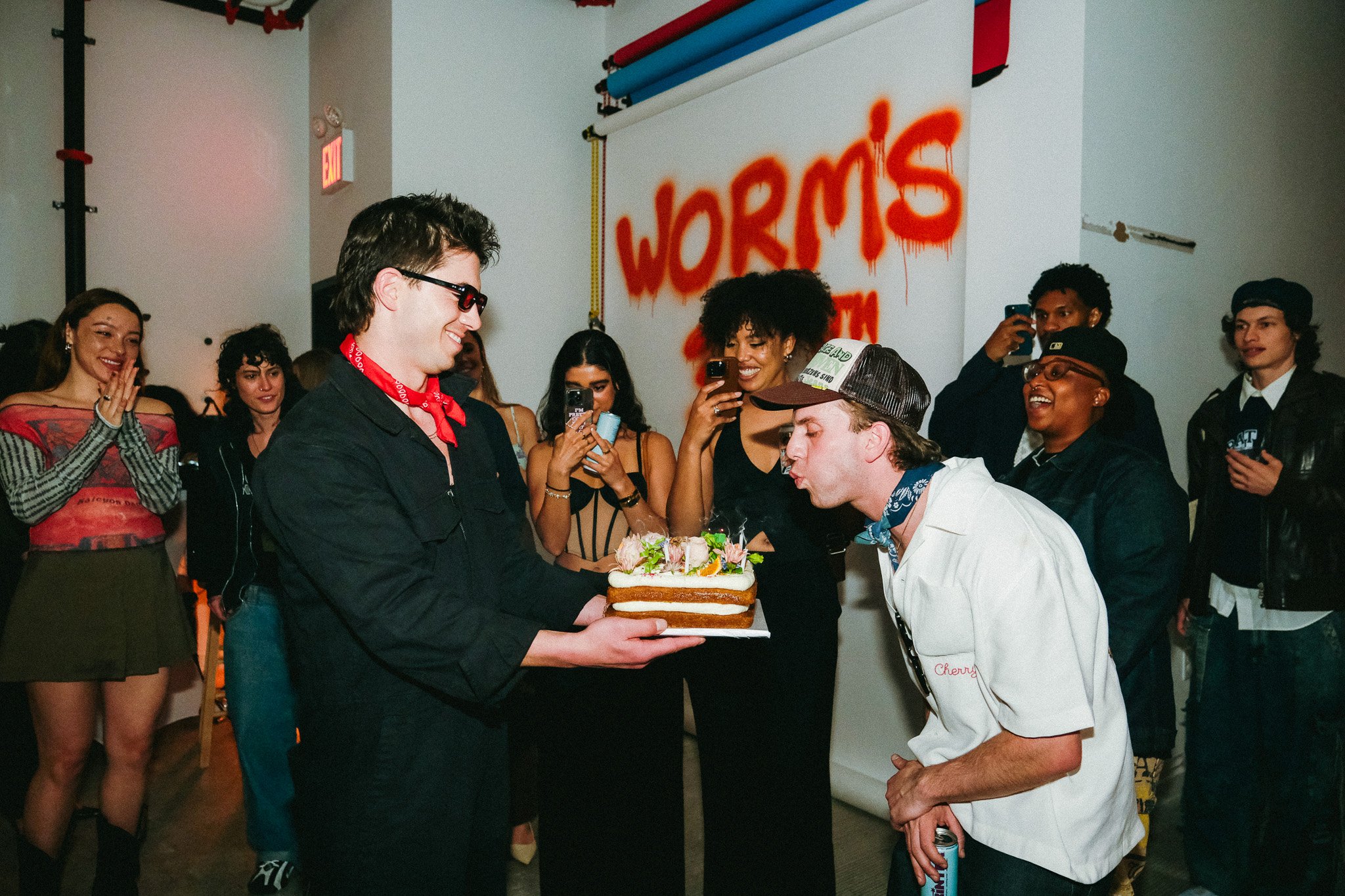 A group of people at a celebration, with one person offering a cake with flowers to another person wearing a white chef's coat, surrounded by lively onlookers.