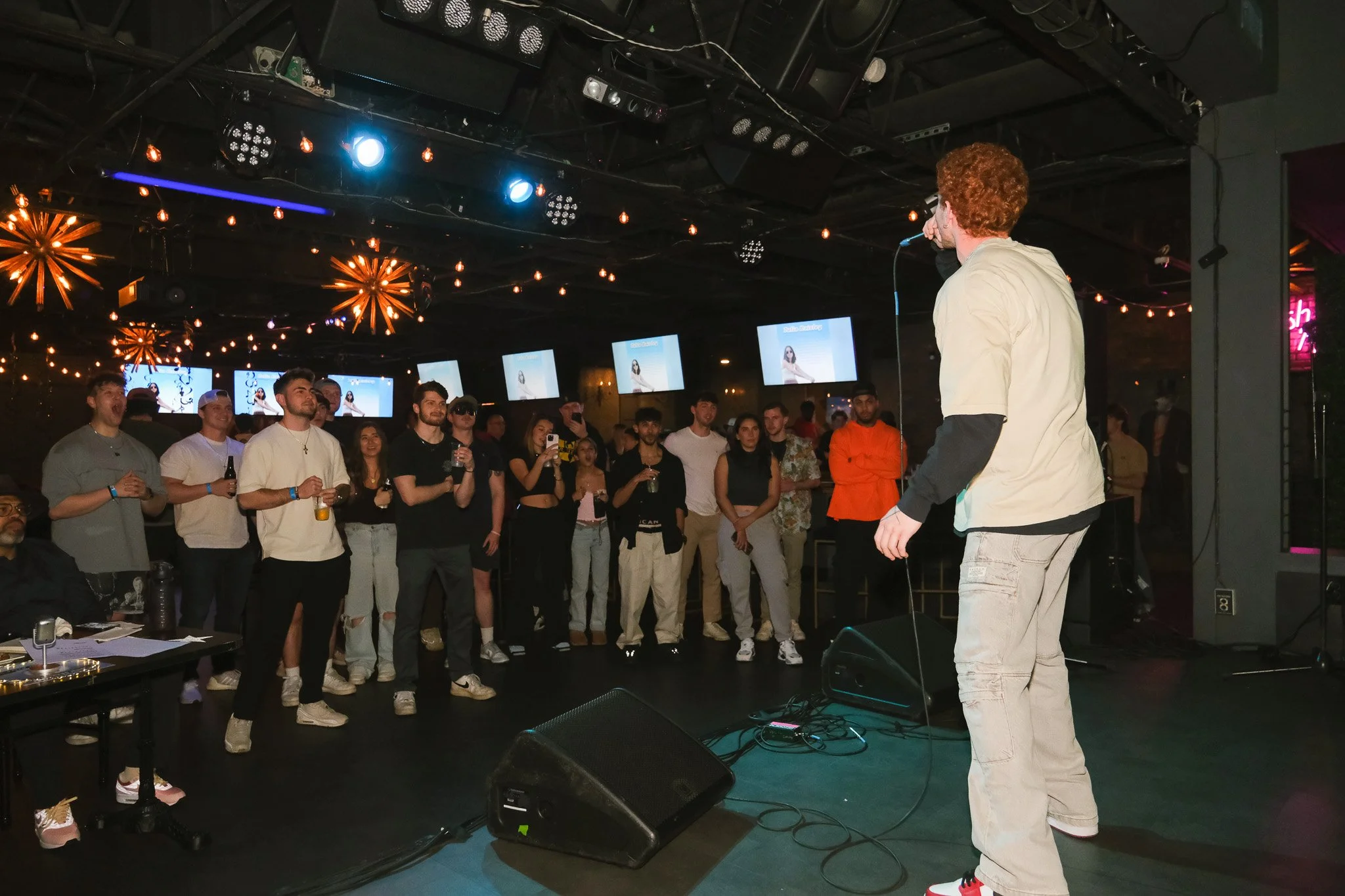A performer on stage with a microphone in hand, facing a crowd of people watching in a dimly lit venue with decorative hanging lights and multiple screens displaying the same female model.