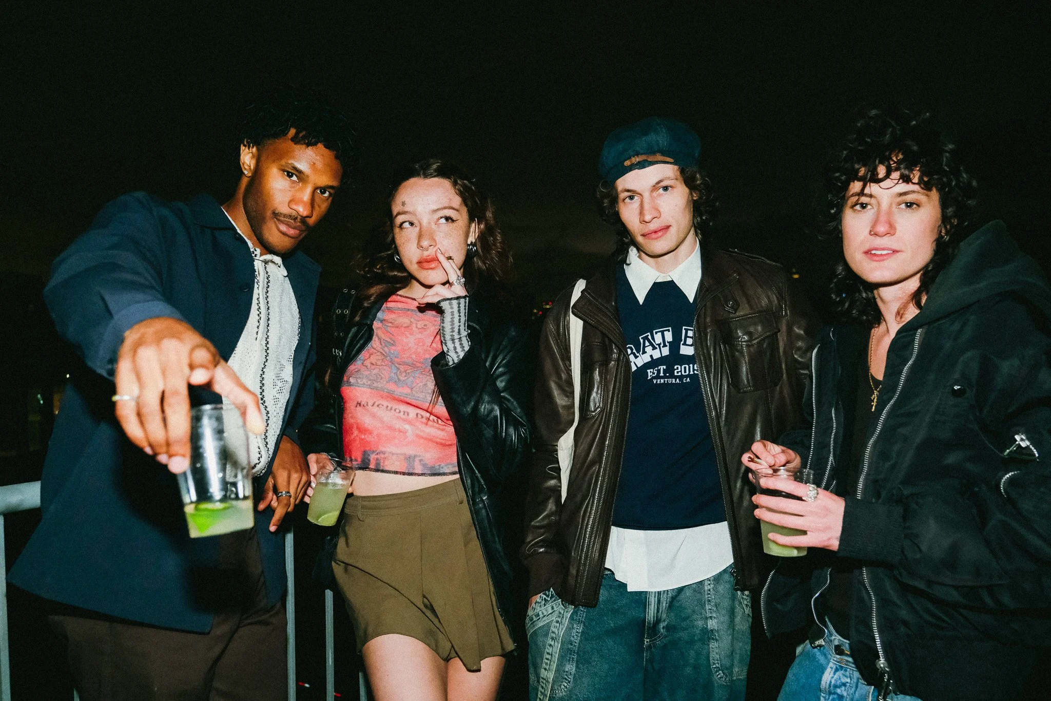 A diverse group of four friends at a nighttime gathering, holding drinks, in casual and stylish clothing, smiling and posing for the camera.