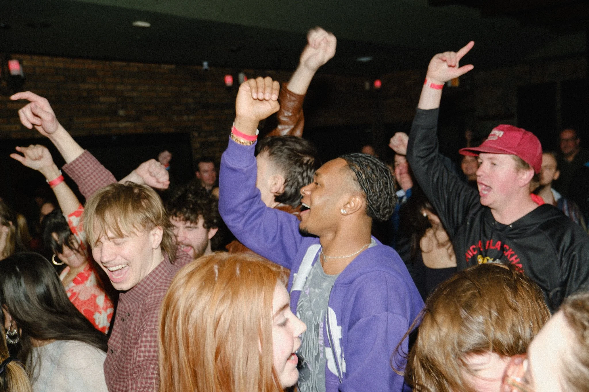 Crowd of people at a concert or event, with many raising their hands and smiling, in a lively indoor setting.