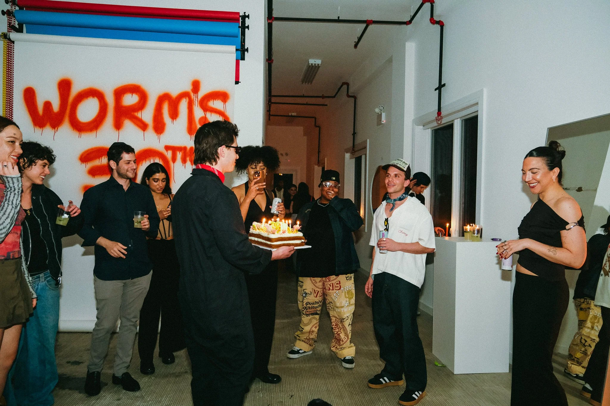 Group of people celebrating a birthday with a cake, candles, and drinks in an indoor space with a large graffiti-style sign on the wall reading 'WORMS'.