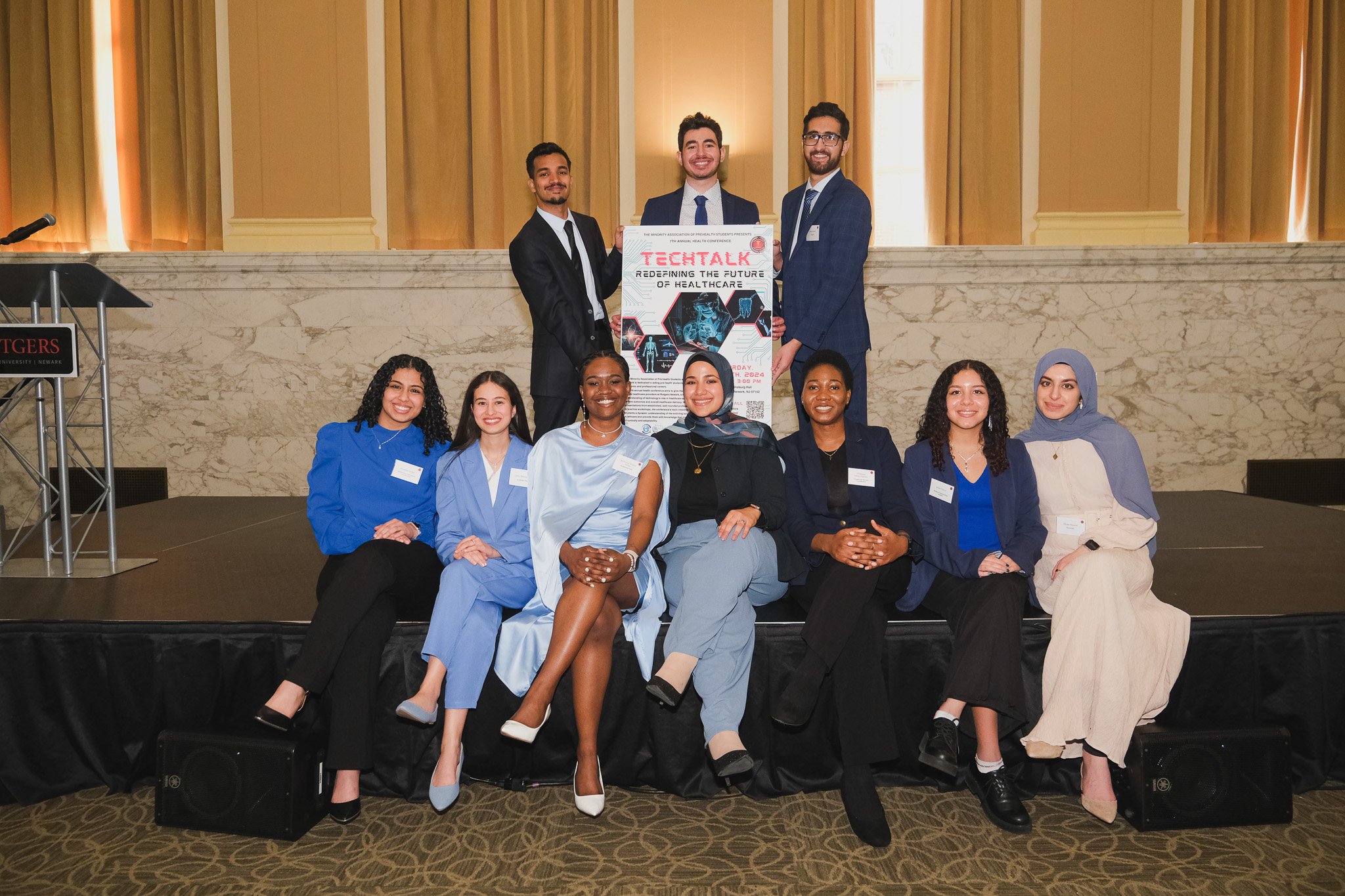 Group of nine young professionals, seven women and two men, posing on a stage with a conference poster that reads 'TechTalk: Redefining the Future of Healthcare' in a conference room with beige walls and golden curtains.