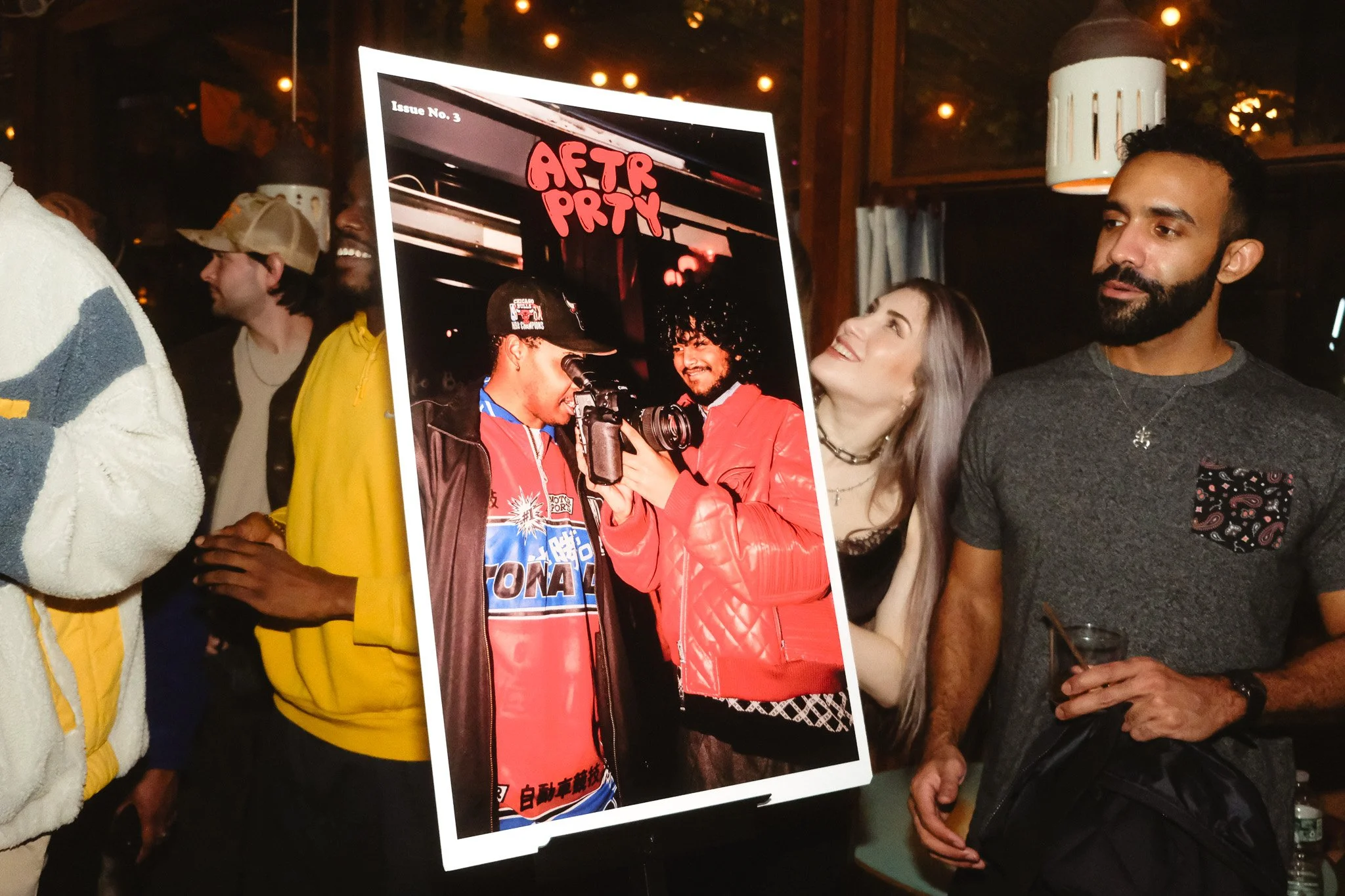 Group of people gathered around a poster of two individuals with a camera, with the word 'AFTR PRTY' written in red on the poster in a dimly lit room.