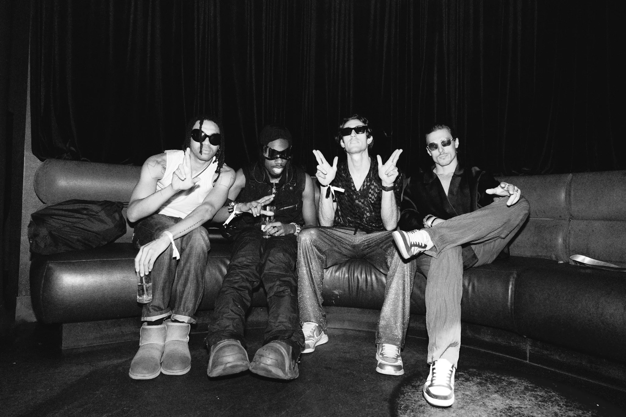 A black and white photo of five young people sitting on a couch in front of a dark backdrop, making peace signs and wearing sunglasses.