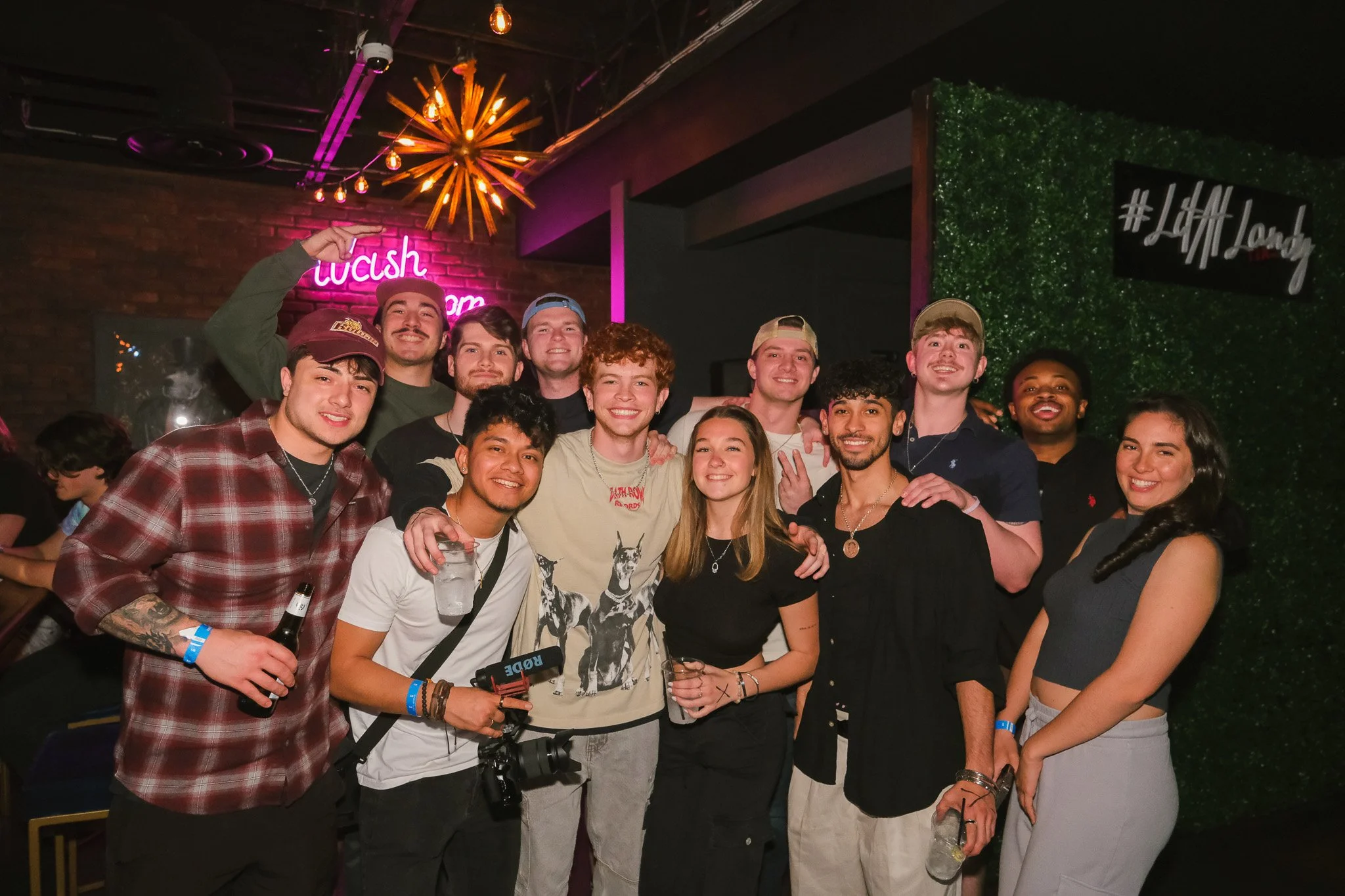 A group of young adults smiling and posing for a photo at a social gathering or party, with some holding drinks, in a dimly lit indoor venue with colorful neon signs and decorative lighting.