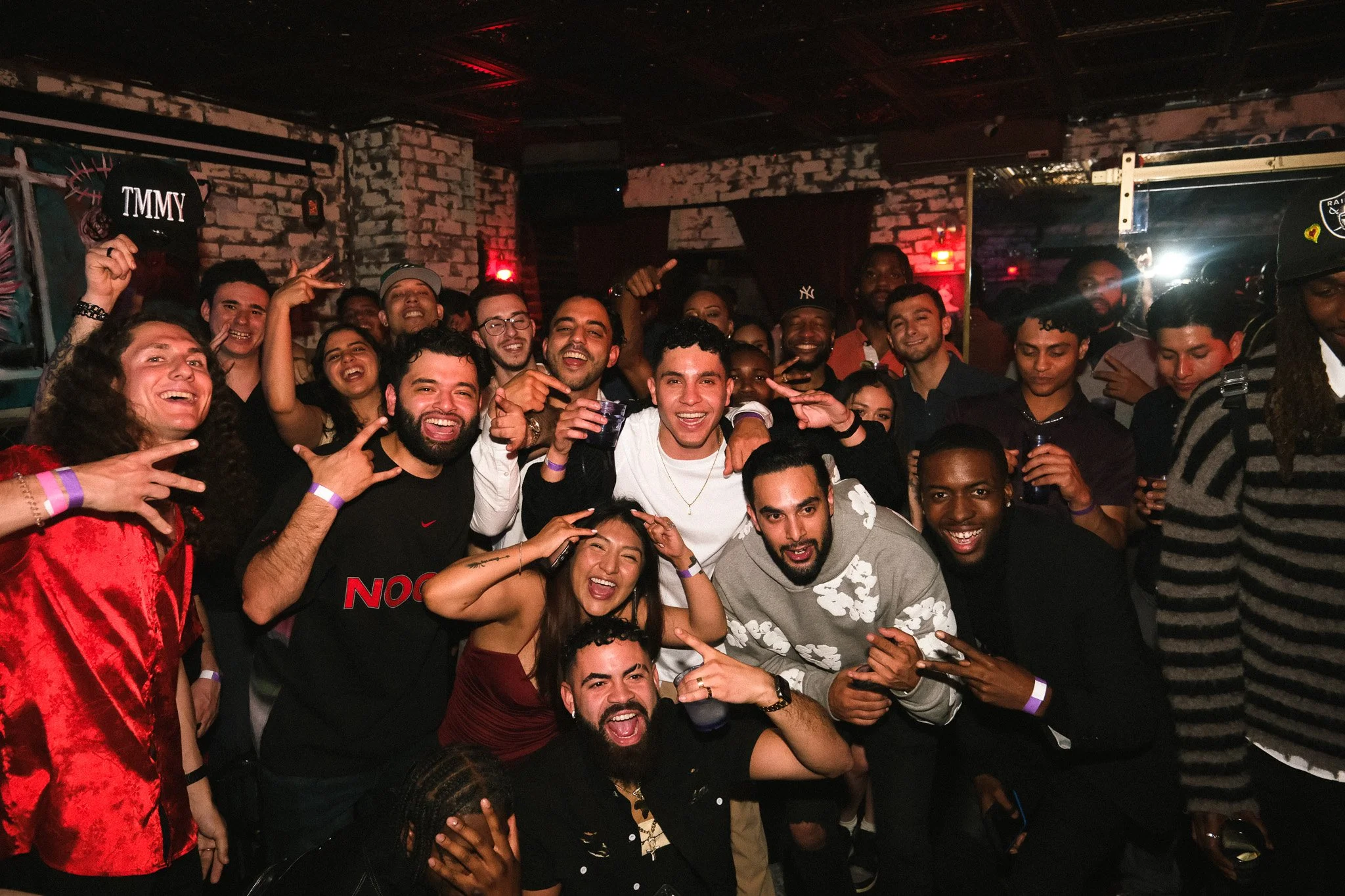 A group of people in a nightclub or bar, smiling and having fun. Some are making peace signs, others are holding drinks, and everyone appears to be enjoying the night.
