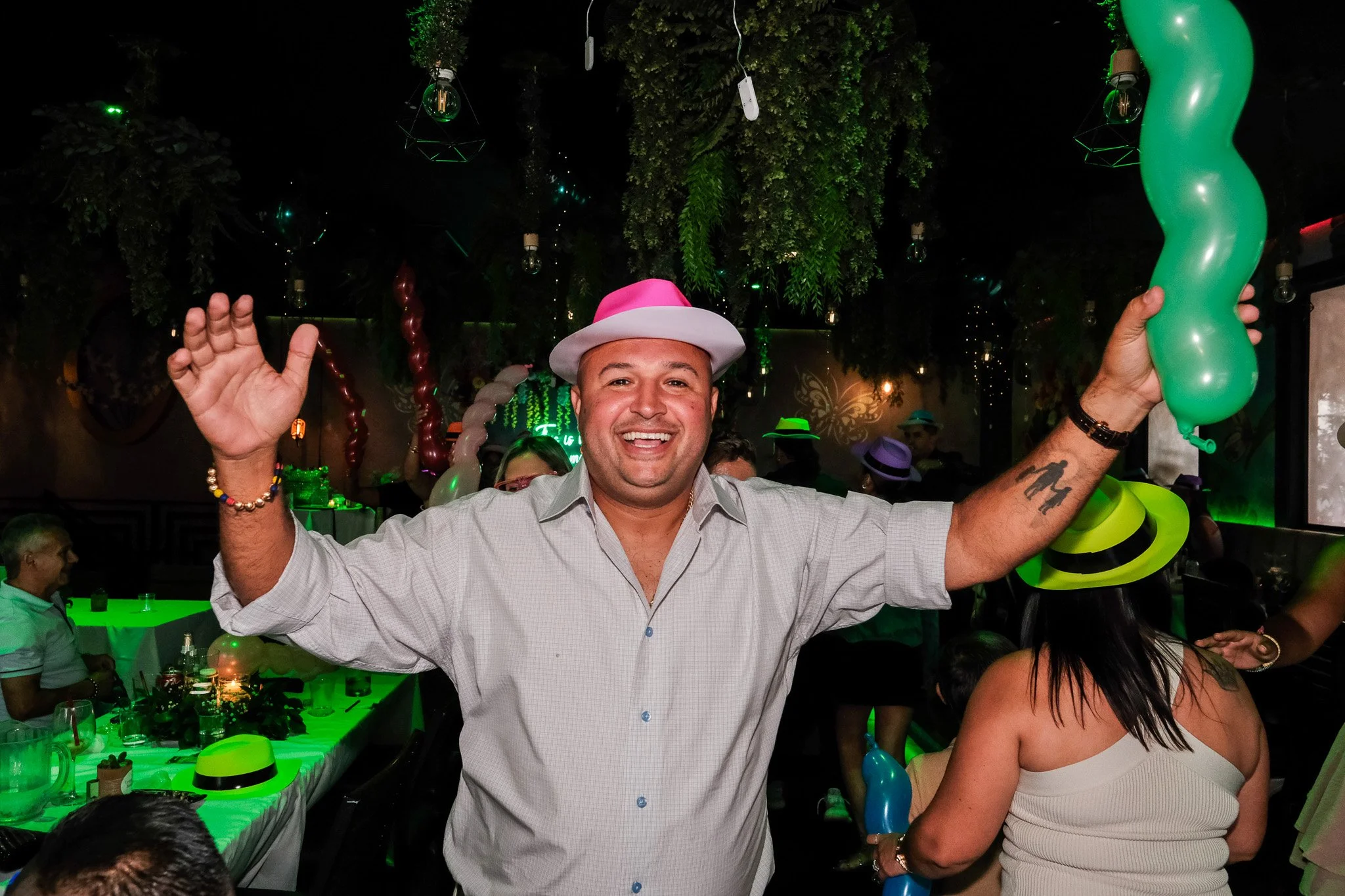 Man wearing a white shirt and pink and white hat smiling and waving at a celebration with green balloons, balloons, and decorations in a festive indoor setting.