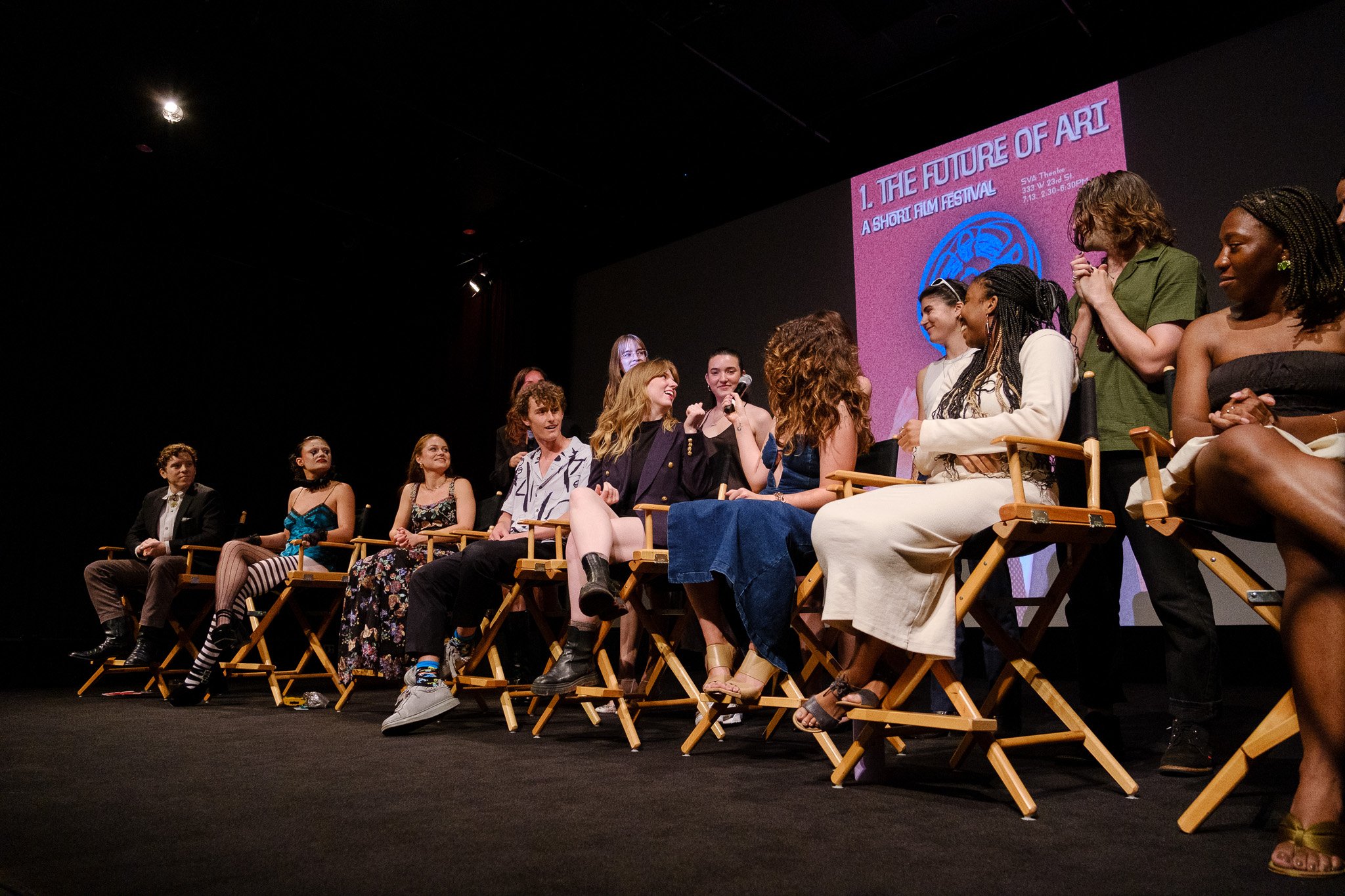 A group of diverse young women and men sitting on stage in wooden chairs during a panel discussion or presentation at an event. A large screen behind them displays text about a short film festival.