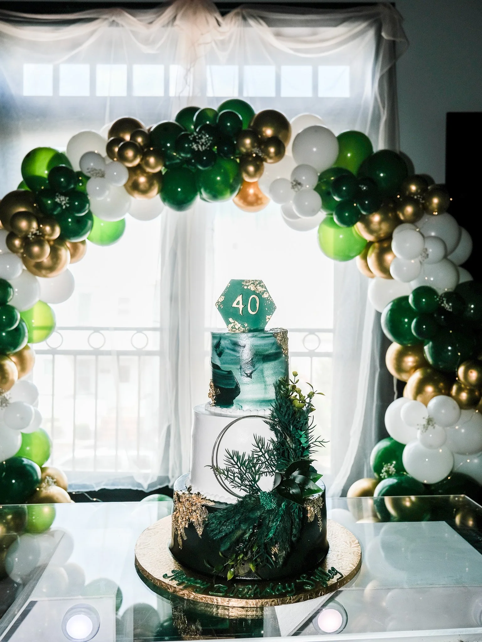 A decorated celebration cake with a nature-inspired design, topped with a green number '40' sign, surrounded by green, white, and gold balloons, set in front of a window with sheer curtains.