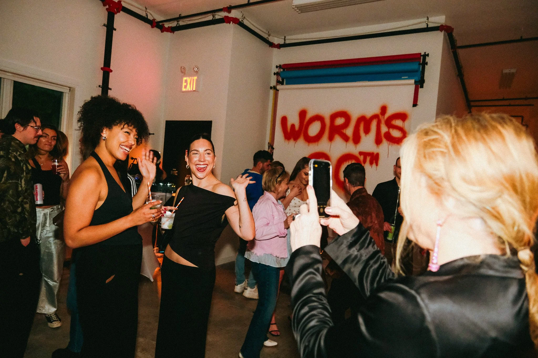 People at a party or social event, smiling and enjoying themselves, with some holding drinks. The setting appears to be indoors with graffiti on the wall that says 'WORMS' and 'COT'.