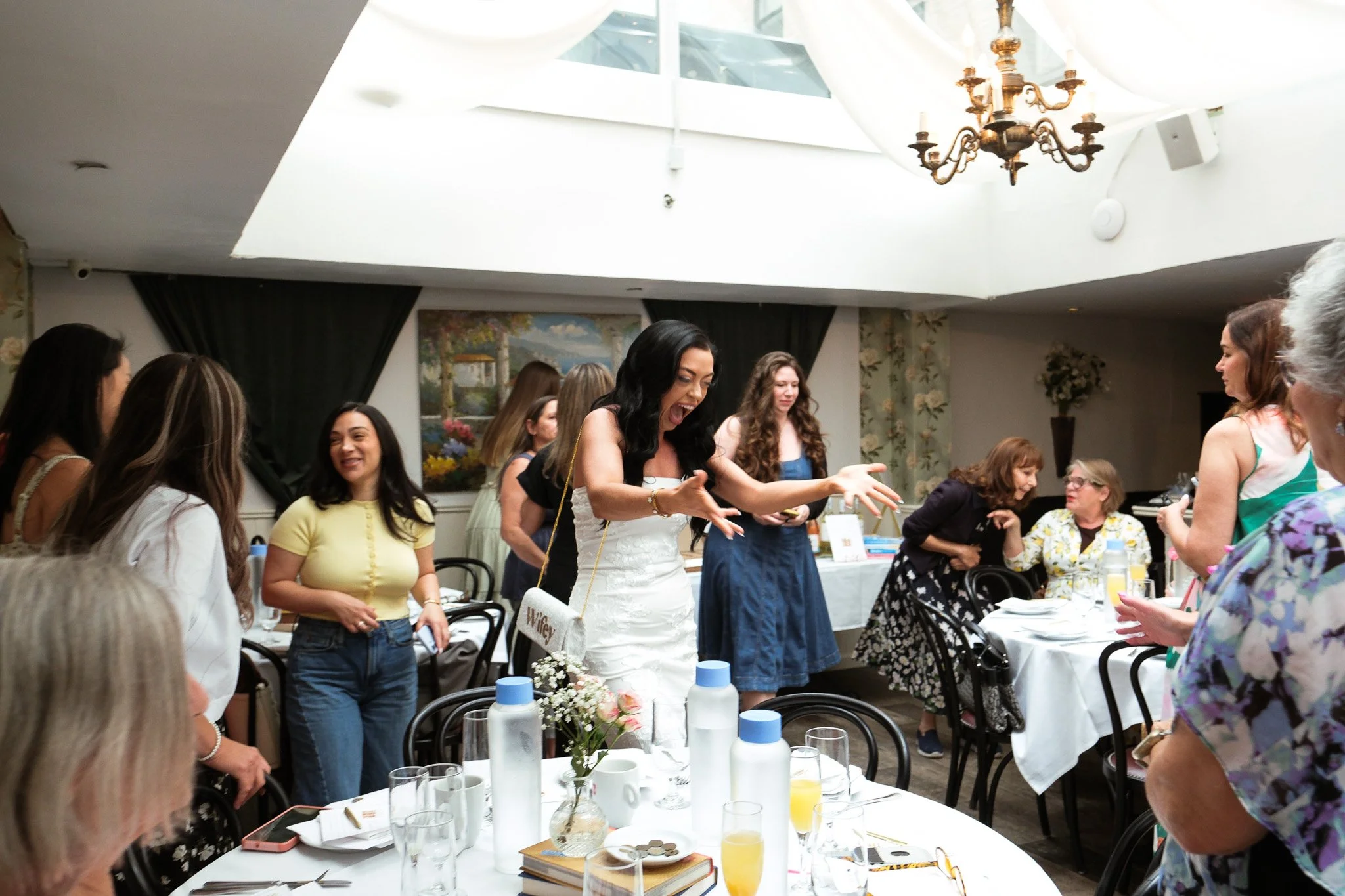 A lively indoor gathering of women around a banquet table, with some standing and engaging in animated conversation. The room has a high ceiling with a large window or skylight, a chandelier, and decorative floral wallpaper.