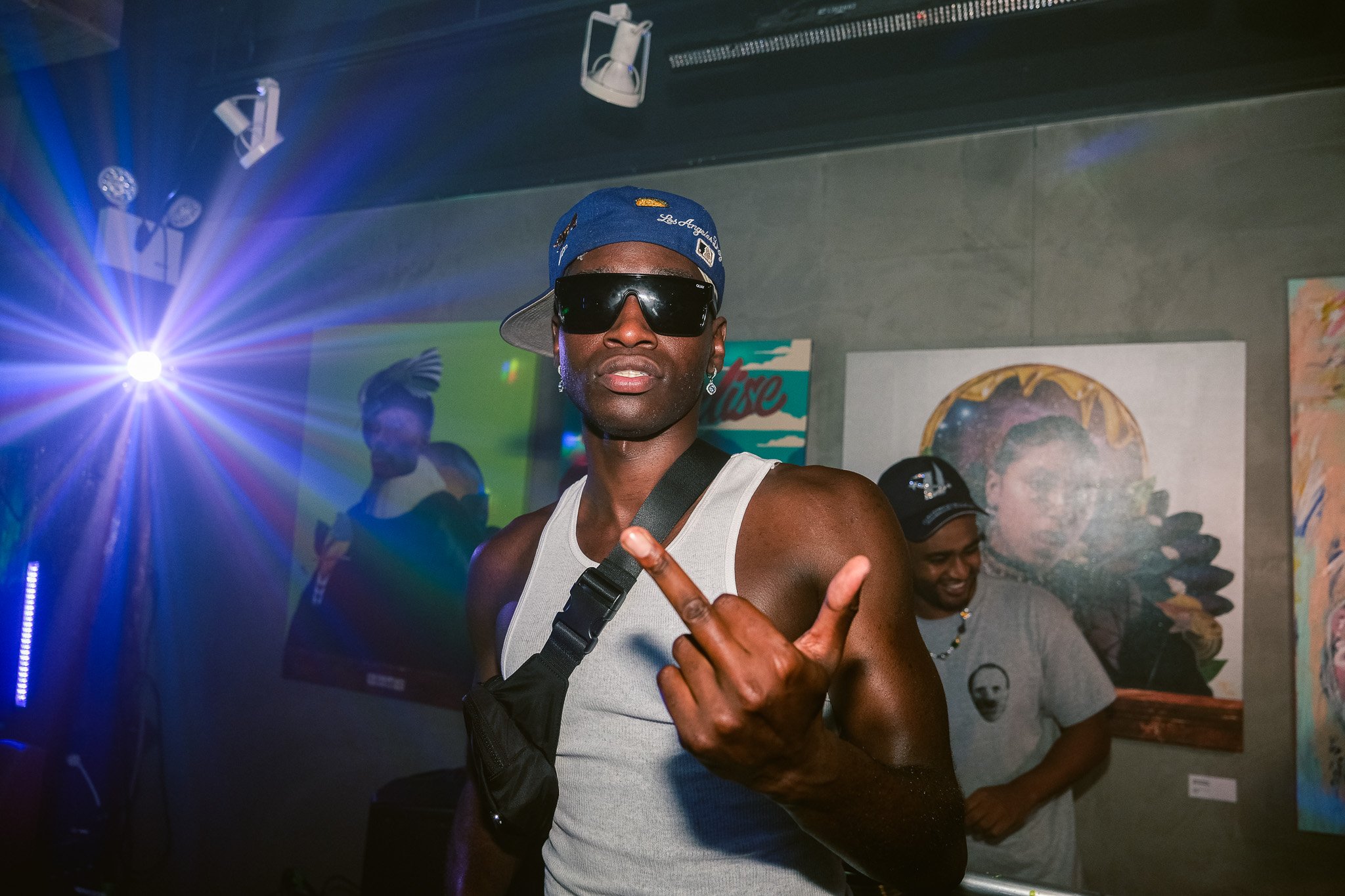 Man wearing a blue Los Angeles Dodgers cap, black sunglasses, and a white tank top, making a hand gesture at a party or nightclub with colorful lights and artworks on the wall behind him.