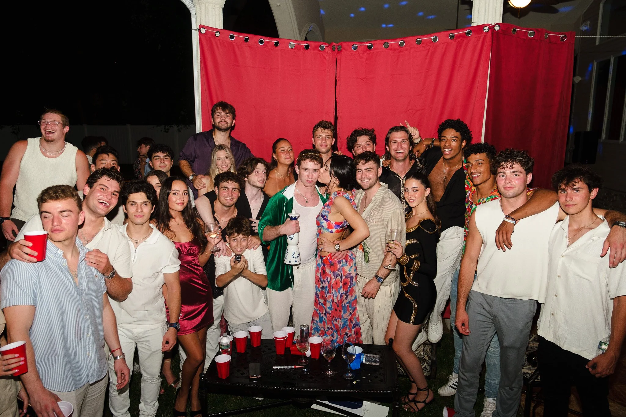 Large group of young adults celebrating at a party outdoors at night, with a red curtain backdrop, holding drinks, smiling, and posing for the photo.