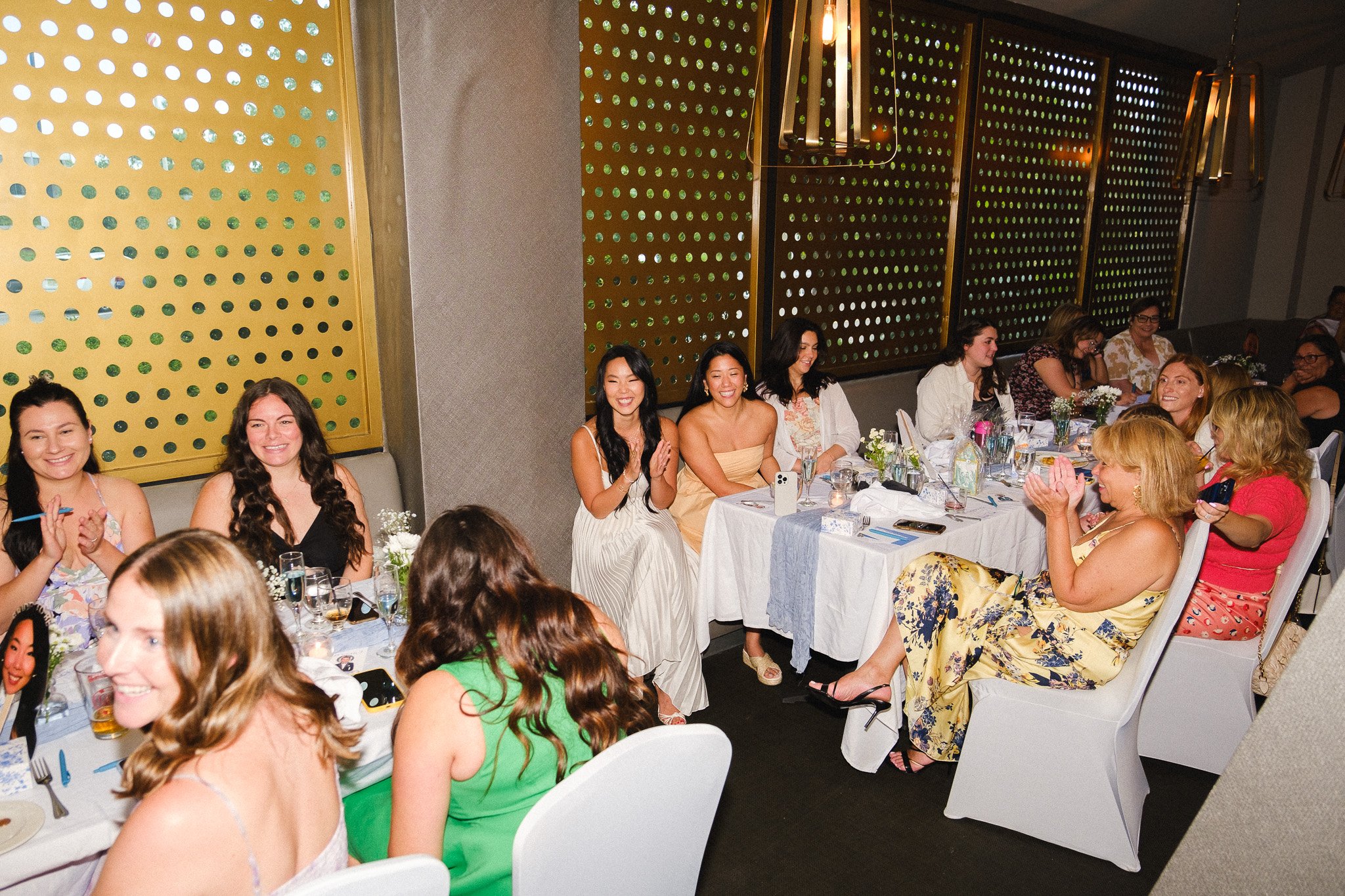 Women gathered around a dining table during a celebration, smiling and clapping, with floral arrangements and glasses on the table in a decorated venue with modern, yellow accent walls.